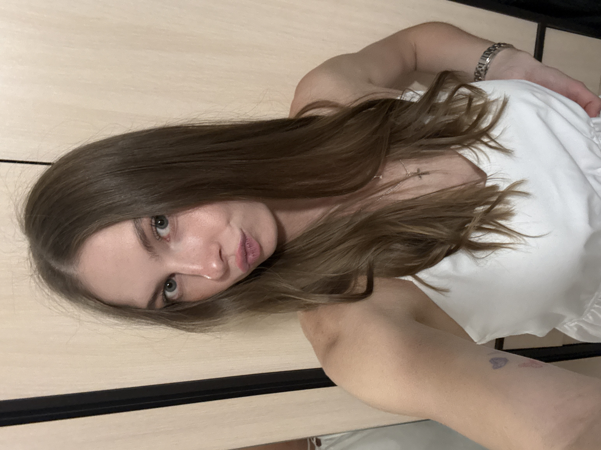 The media file features a young woman lying down, with her head tilted to the side. She has long, brown hair and is wearing a white top. A silver bracelet is visible on her wrist, and a faint tattoo, possibly a heart shape, is on her arm. Her eyes are open and she is looking towards the camera. Her lips are slightly parted, and she appears to be wearing lipstick. The background is a light-colored, wooden surface, possibly a table or a wall. There is a dark horizontal line across the image, suggesting a seam or a border. The lighting appears to be soft and diffused, indicating it might be daytime or an indoor setting with artificial light. The overall mood is relaxed and intimate. The location is given as Perm, Russia.