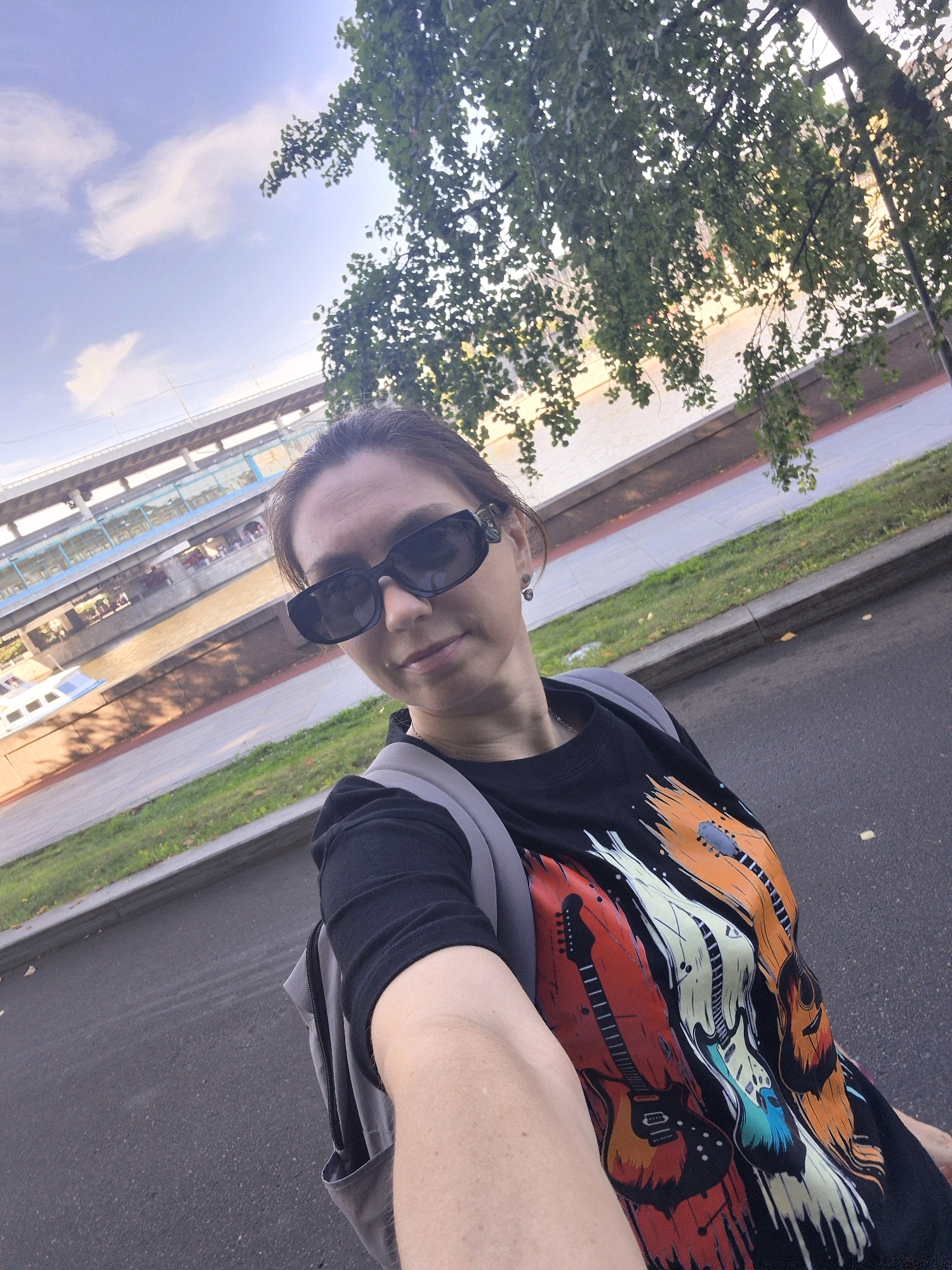 A woman with dark hair, wearing sunglasses, a black t-shirt with a colorful guitar print, and a grey backpack, stands on a paved surface. In the background, there is a body of water with a boat, and a multi-level structure that appears to be a bridge or elevated walkway. The scene suggests an urban environment, potentially a waterfront area in Moscow, Russia, given the architectural style and natural elements. The lighting indicates it is daytime, with sunlight casting shadows.