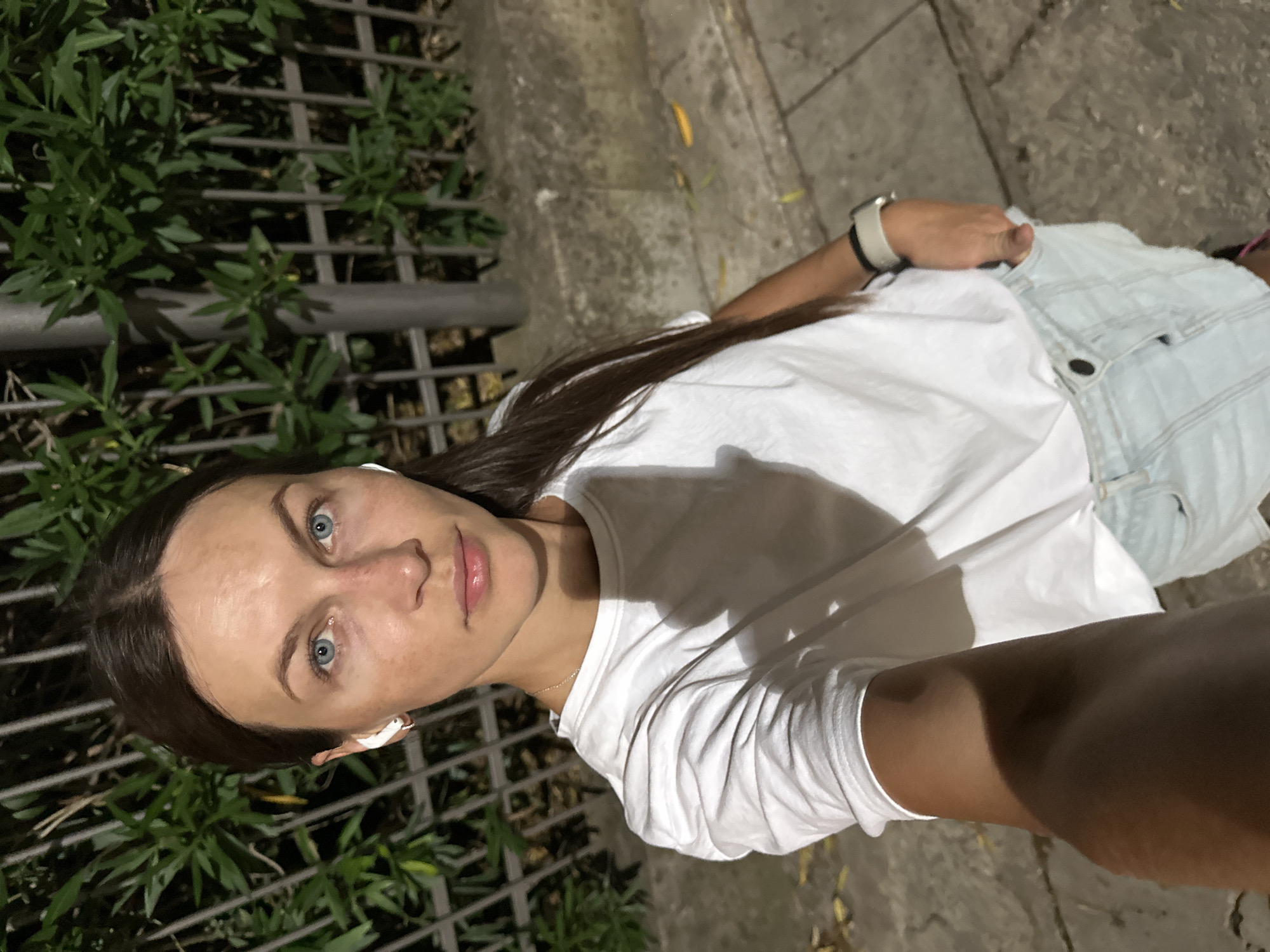 A woman stands outdoors at night, taking a selfie. She is wearing a white t-shirt and light blue denim shorts. She has earbuds in her ears and a watch on her left wrist. Behind her, there is a metal fence and green foliage, and a concrete wall and pathway. The location is in the city of Boreti, Montenegro.