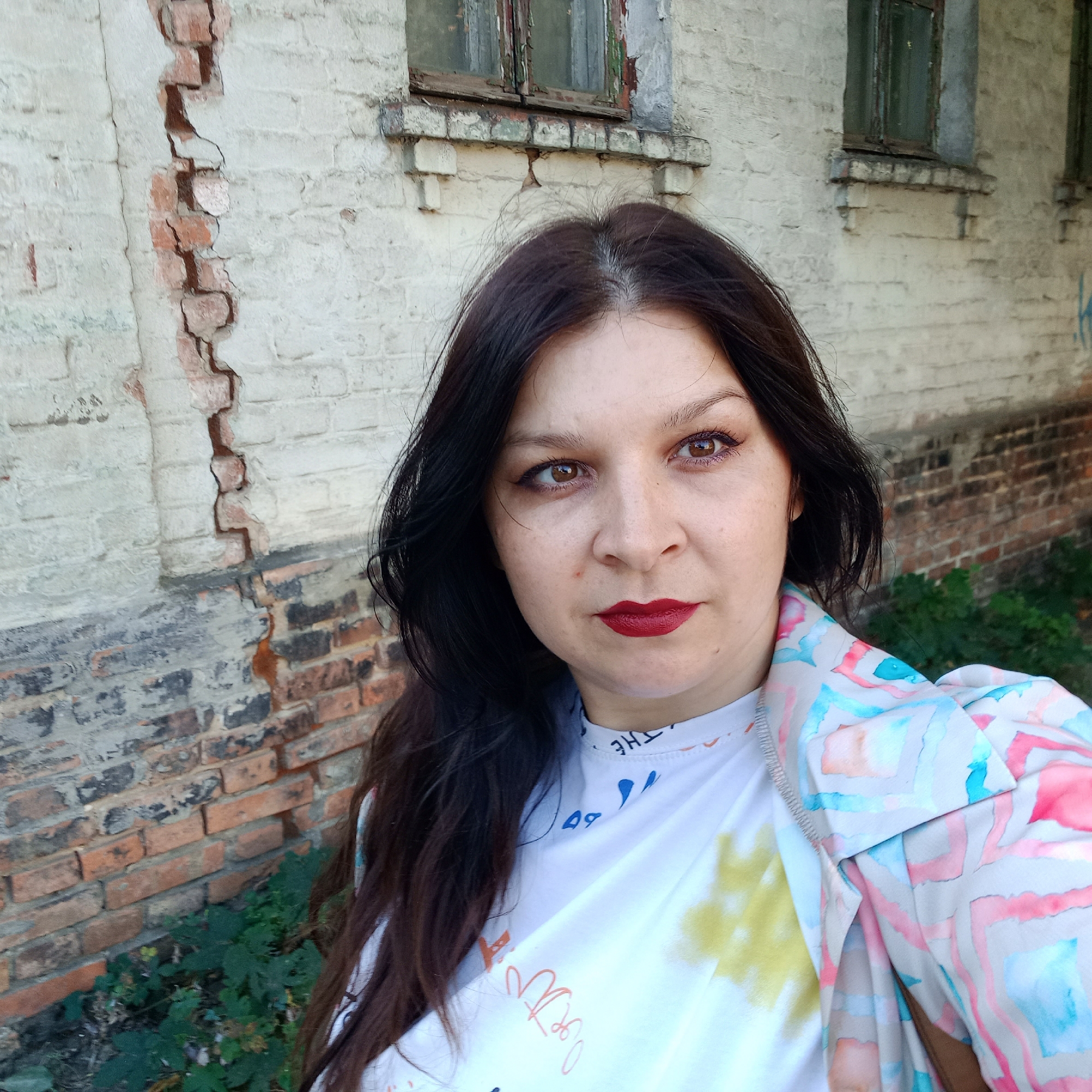 The image shows a woman in a selfie taken outdoors. She has dark brown hair and is wearing dark lipstick. She is also wearing a white t-shirt and a jacket with a colorful, abstract pattern. The background is a distressed brick building with peeling paint on the windows. The building appears to be old and somewhat neglected. The lighting suggests it is daytime. The scene is likely located in an urban or semi-urban environment. Kropyvnytskyi, Ukraine is a city with a mix of older architecture and more modern structures, and this image is consistent with such a setting.