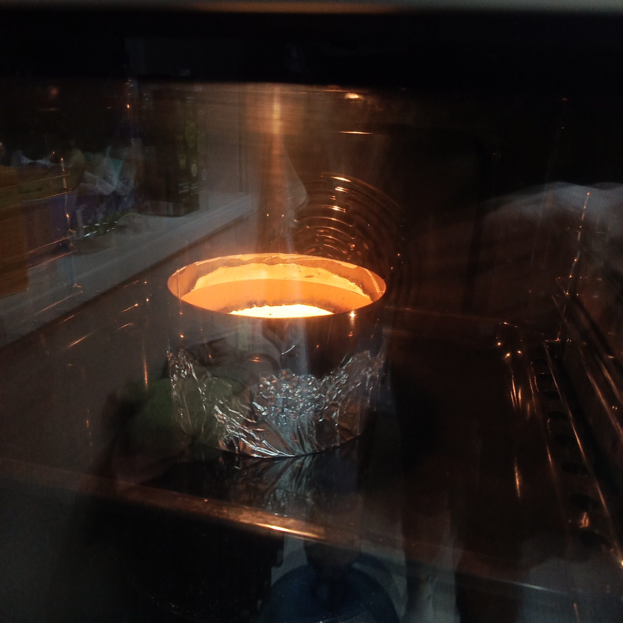 Inside an oven at a residence in the city of Марковский, Russia, a silver, round baking pan is positioned on an oven rack. The lower portion of the pan is wrapped in crinkled aluminum foil. The pan contains a light-colored, possibly baking, mixture that radiates an orange glow from the oven's internal heat source. The dark interior of the oven reflects ambient light and indistinct shapes, suggesting a room environment outside the oven's glass door. No individuals are present in the image.