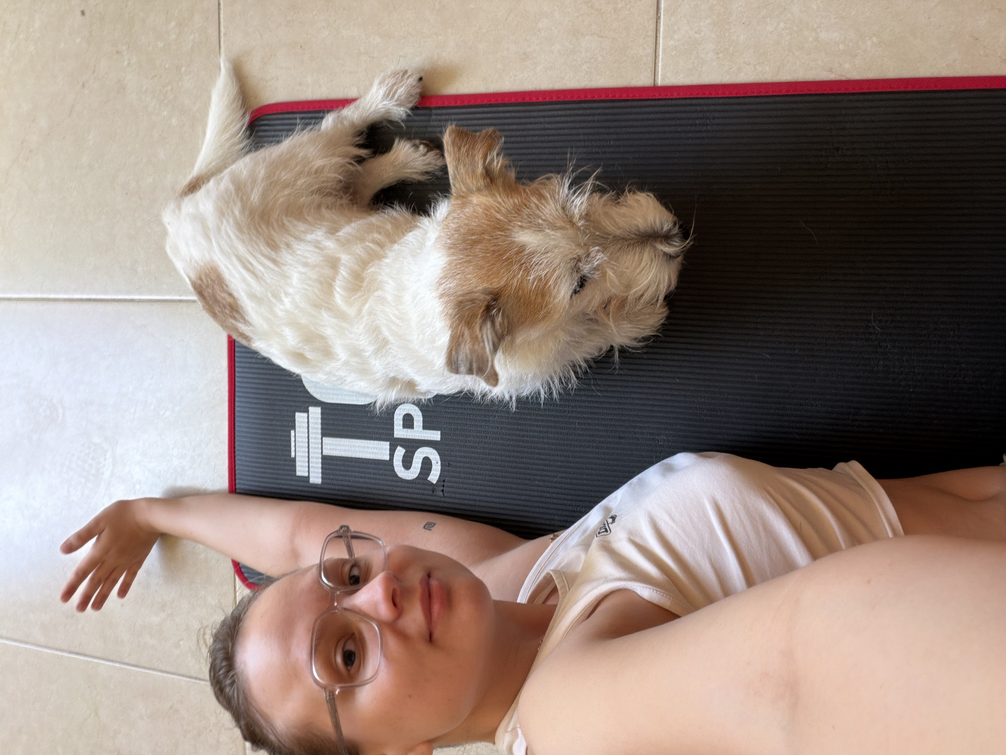 An overhead shot captures a woman and a small dog relaxing together on an exercise mat, likely indoors at a home in Tel-Aviv, Israel.
The main subject is a woman with light brown hair, wearing clear-framed glasses and a light-colored tank top. She is lying on her back on a black exercise mat, extending one arm to the side, possibly holding the camera for a selfie. She looks directly towards the camera with a neutral, calm expression.
Alongside her, a small dog with scruffy white and brown fur is also lying down on the mat, appearing very relaxed. Its head is turned away from the camera, and its body is partially curled.
The mat itself is black with a distinctive red border. White graphics are visible on the mat, including a stylized icon resembling a weightlifting dumbbell or barbell, and the partially visible text "SP" followed by "TS", suggesting the word "SPORTS". The surrounding floor is made of light-colored, possibly beige or cream, tiles with visible grout lines.
The lighting is soft and natural, indicating an indoor daytime setting. The overall mood of the image is peaceful and domestic, showing a moment of quiet companionship between the person and her pet.