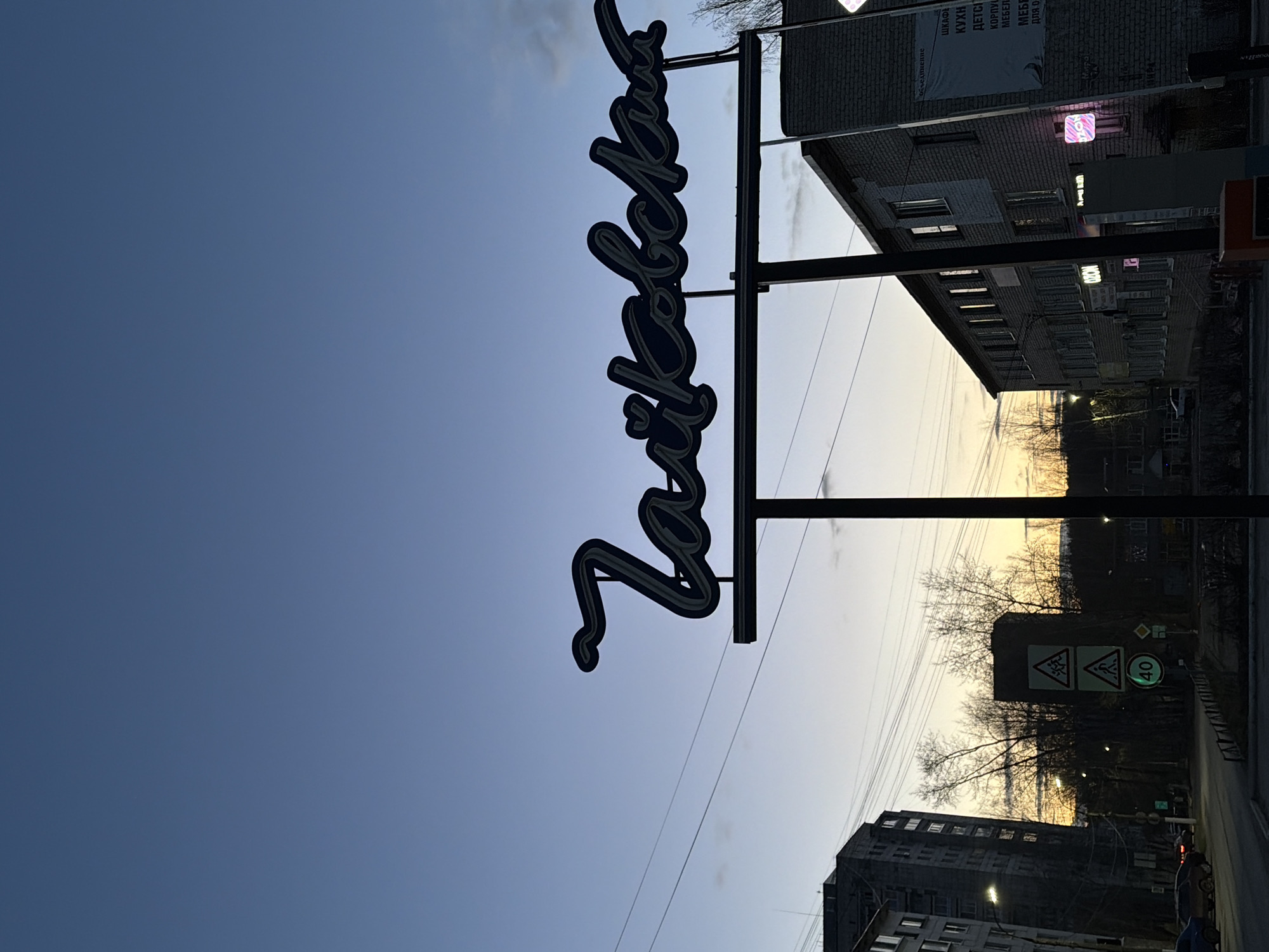 This media file depicts an urban street scene at either dawn or dusk, set against a clear, transitioning sky from deep blue overhead to a lighter, yellowish hue near the horizon. The image is composed in a portrait orientation, with the sky occupying the majority of the left side.
A prominent feature is a large, dark, cursive neon sign, suspended from a tall pole, which reads "Заводчанка" (Zavodchanka) in Cyrillic. This sign is silhouetted against the sky. To the right, several multi-story buildings, characteristic of post-Soviet architecture, line the street. One building displays a large banner with Russian text: "ШКАФЫ КУХНИ ДЕТСКИЕ МЕБЕЛЬ ДЛЯ ДОМА" (Wardrobes, Kitchens, Children's, Furniture for Home) and below it, "объединение" (association). Several illuminated neon signs with a pinkish glow are visible on the building facades, suggesting commercial establishments.
Bare trees, indicative of autumn or winter, are silhouetted against the lighter part of the sky in the background, flanking what appears to be a road or street. Traffic signs are also visible; one triangular sign depicts two figures (likely a children crossing warning), and a circular sign shows "40", indicating a speed limit. Power lines stretch across the frame. The overall atmosphere is quiet and still, emphasizing the urban environment at the transition of day and night. The location context indicates this scene is in Chaikovsky, Russia.