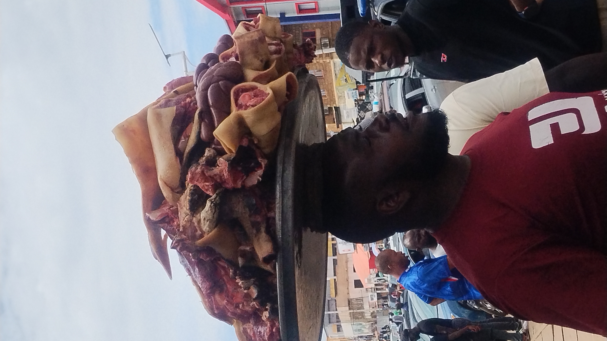An outdoor urban scene in Jos, Nigeria, captures a bearded man in the foreground carrying a large, round, flat metal tray on his head. The tray is heavily laden with a diverse assortment of raw meat and offal, including reddish cuts of muscle tissue, light beige rolled animal skin or tripe showing exposed flesh, and dark, purplish, convoluted organs such as intestines or stomach. The man is wearing a red t-shirt with a white graphic design and is oriented facing the right side of the frame. To his right, another man in a black t-shirt with a red logo is visible, looking towards the left. In the mid-ground and background, a street scene unfolds with numerous parked cars. Several other individuals are present, including a man in a blue t-shirt further to the left. Multi-story buildings with varied facades, including a prominent red and white structure on the right, line the street. The sky is overcast with light grey clouds.