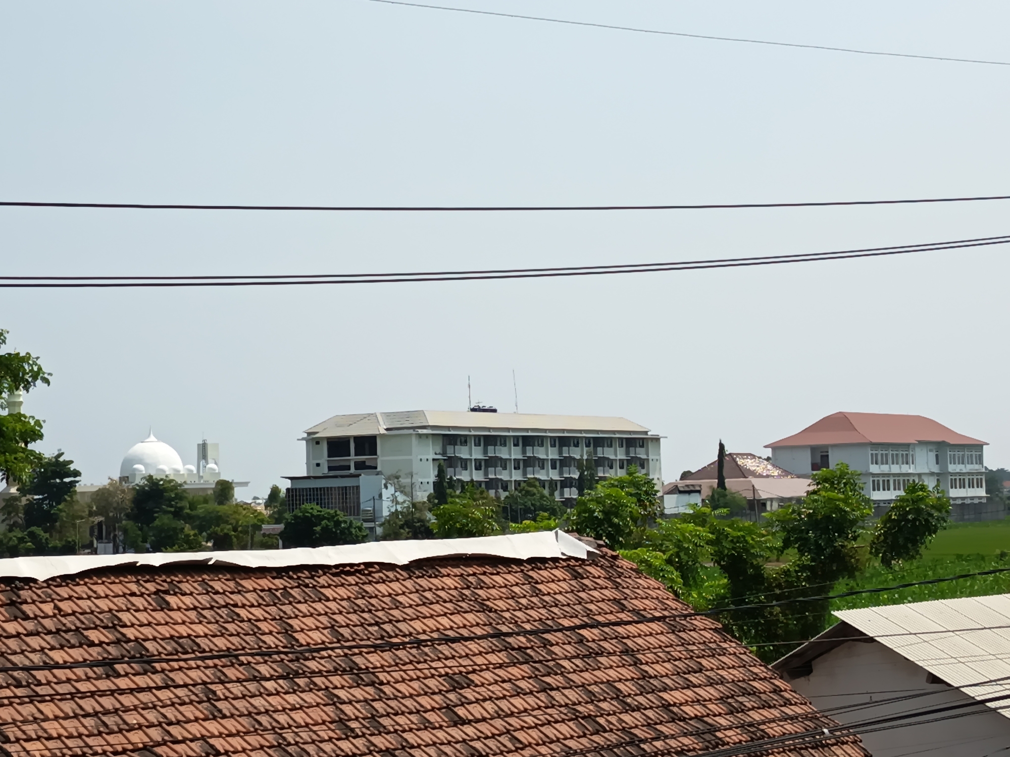 The image depicts a view of a residential area in Gebangan, Indonesia, with several buildings and lush greenery under a clear sky. In the foreground, the terracotta tiled roof of a house dominates the lower portion of the frame. Beyond this, a large, light-colored multi-story building with many windows is visible, suggesting it might be an apartment complex or an office building. To the left of this building, a white dome, likely part of a mosque, stands out against the sky. Further to the right, another multi-story building with a reddish-brown roof is situated. The middle ground is filled with dense green trees and foliage, creating a natural buffer between the foreground and the distant structures. The background consists of a hazy sky with power lines crisscrossing the upper part of the image. The scene appears to be captured during daylight hours. There are no people or animals visible in the image, and no specific actions or interactions are depicted. The environment is characterized by a mix of residential buildings, natural vegetation, and overhead electrical infrastructure, typical of an urban or semi-urban setting in Indonesia.