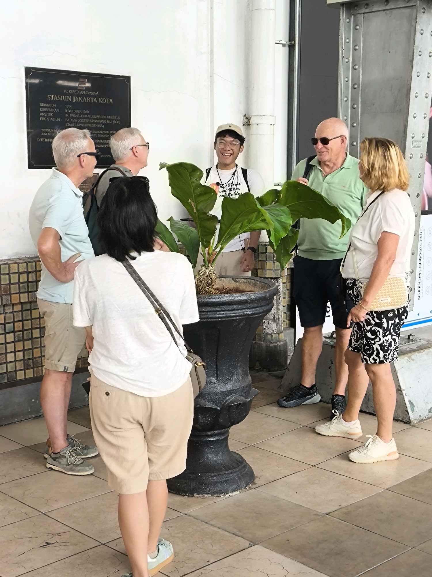 In West Jakarta, Indonesia, a group of five individuals is gathered around a large potted plant in front of a building with a historical plaque. The plaque, visible on the left, is black with white text and indicates "STASIUN JAKARTA KOTA" (Jakarta Kota Station), built in 1914.
Two older men in casual attire, one in a light blue polo shirt and khaki shorts, the other in a green polo shirt and dark shorts, are conversing. The man in the blue shirt is facing away from the camera, while the man in the green shirt is facing partially towards the camera, wearing sunglasses. A blonde woman in a white top and a black and white patterned skirt stands next to the man in the green shirt, looking towards him.
A young man wearing glasses and a light-colored baseball cap stands to the right of the plaque, smiling and looking towards the other individuals. He is wearing a white t-shirt with text and a backpack. In the foreground, a woman with dark hair, wearing a white t-shirt and beige shorts, is facing away from the camera and walking.
The environment features a tiled floor and a textured wall. A large, dark pot contains a vibrant green plant with large leaves. The lighting suggests an outdoor or semi-outdoor setting during the daytime.