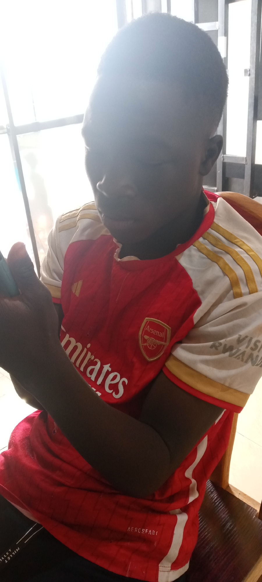 A young person sits indoors, wearing an Arsenal football club jersey. The jersey is red with white sleeves and gold trim, displaying the Emirates sponsor logo. The person is holding a mobile phone in their left hand. The background shows a window with visible sunlight. The location is likely Jos, Nigeria, based on potential information about the image's origin.