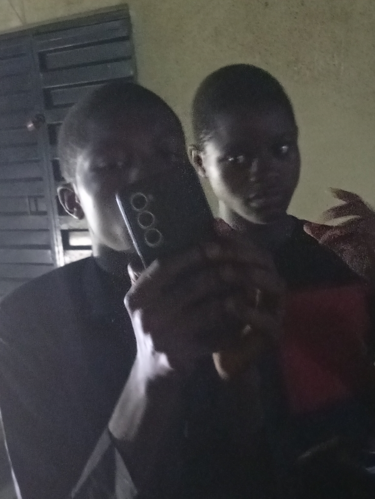 The image shows two individuals in what appears to be an indoor setting. One person is holding a mobile phone with three circular lenses and is partially obscuring their face with it. The other person is standing behind, slightly out of focus. The lighting is dim, and the background is nondescript. A window with metal bars is visible behind one of the individuals. There is no specific evidence to indicate the location as Shangisha, Nigeria.
