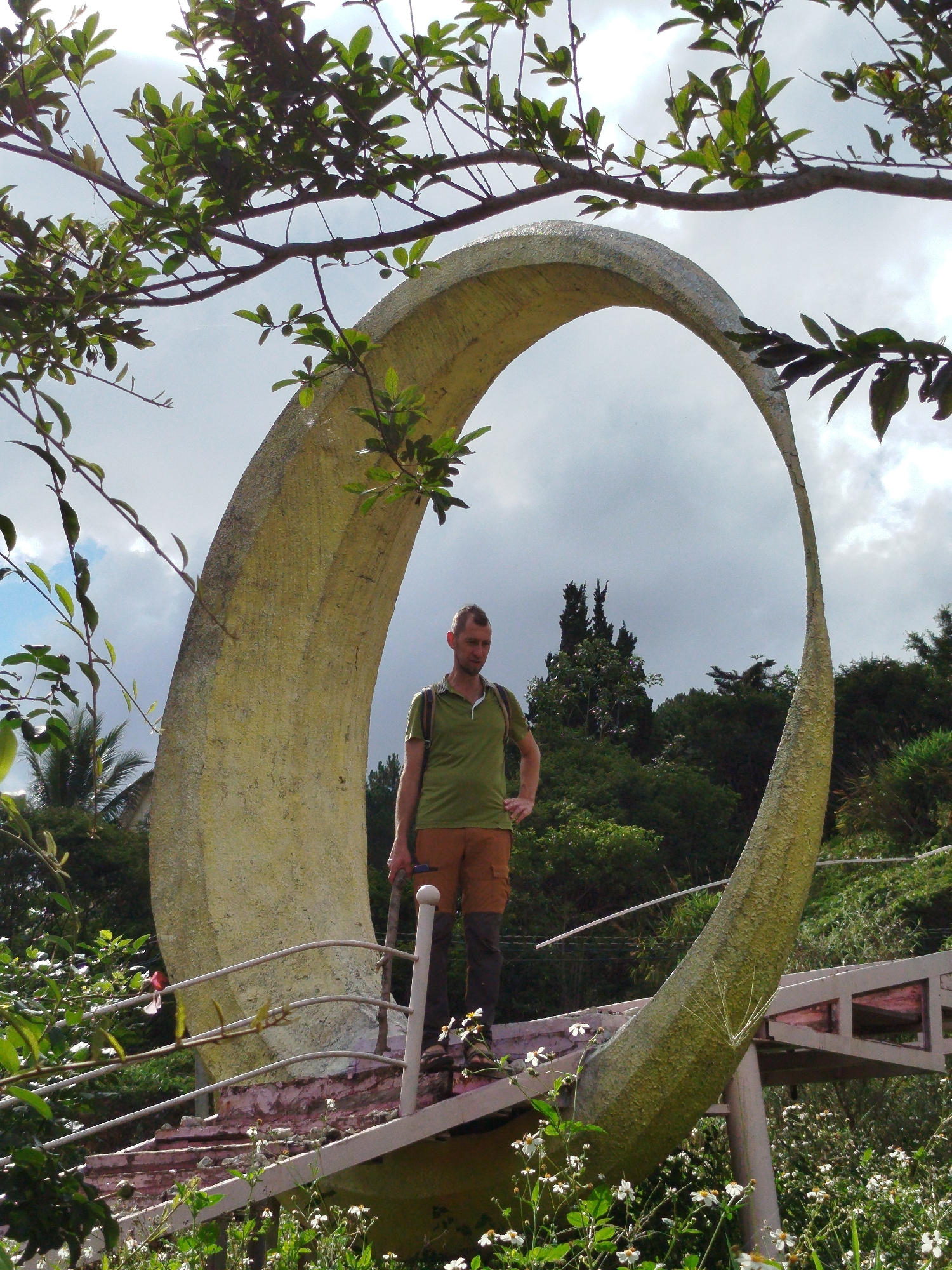 A man stands on a structure holding a walking stick, framed by a large, circular arch. The man is wearing a green polo shirt, brown shorts, and dark gaiters over hiking boots. He is fair-skinned with short hair. The circular arch, constructed of a light-yellow, textured material, is positioned on what appears to be a bridge or viewing platform with white railings. The platform’s steps are visible beneath.
The background features a lush green landscape, consisting of various trees and foliage, beneath a cloudy sky. Branches from surrounding trees arch into the frame at the top and sides. Small white flowers are visible in the foreground vegetation.
This image depicts a scene in Da Lat, Vietnam. The architecture is specific to that region.