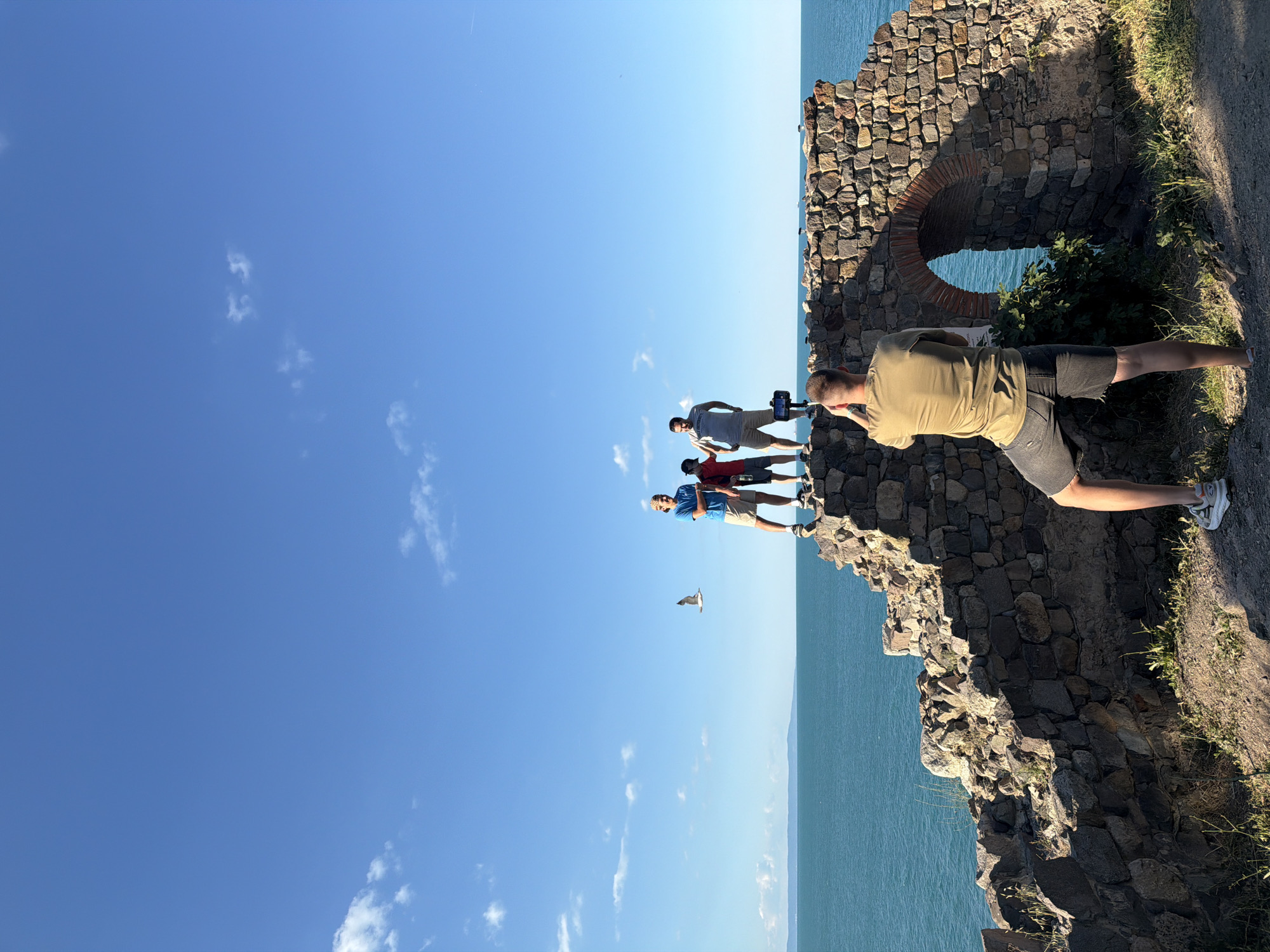 In Sozopol, Bulgaria, a man in a tan t-shirt and dark shorts stands facing away from the camera, with his right arm raised as if taking a photo or video with a phone mounted on a tripod. In front of him, on top of a crumbling stone structure, three people are posing. To the left, a person in a blue t-shirt and beige shorts stands with their left hand on their hip and their right arm outstretched towards the person taking the photo. Next to them, a child in a red t-shirt and dark shorts looks towards the camera. To the right, a man in a gray t-shirt with "BUL UNIVERSITY" text and khaki shorts stands with his hands on his hips. A seagull is captured mid-flight between the posing group and the photographer. The background features a wide expanse of bright blue sea under a clear blue sky with scattered white clouds. The stone structure itself is ancient and weathered, with an arched opening visible on the right side. The ground in the foreground is a mix of dirt and sparse vegetation.