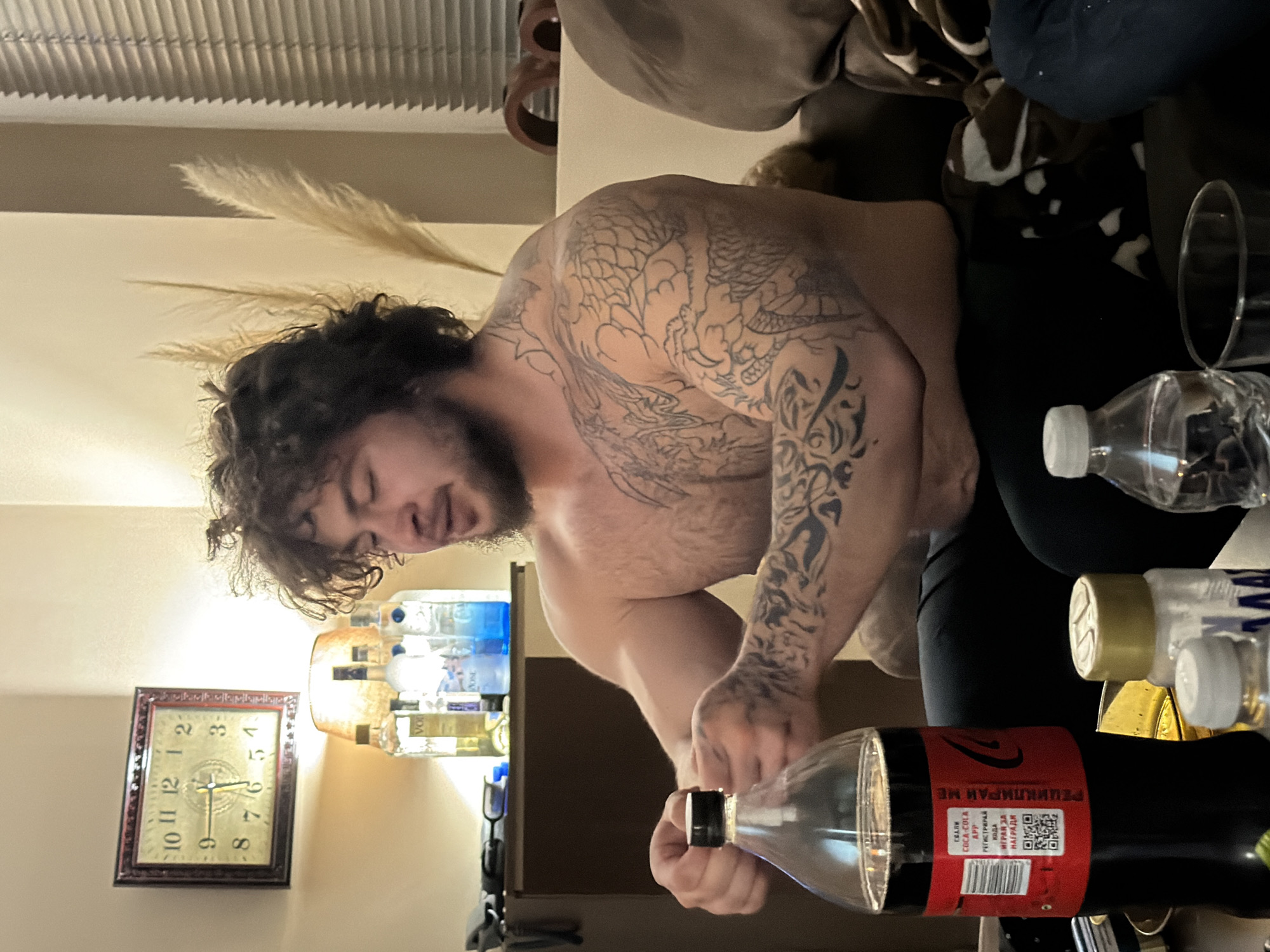 A man with tattoos is opening a bottle of Coca-Cola. He is shirtless, and the tattoos cover his arms and upper back. There is a clock on the wall indicating the time of 11:08, and several bottles of water and alcohol are visible on a nearby shelf. The location and city are not apparent from the image.