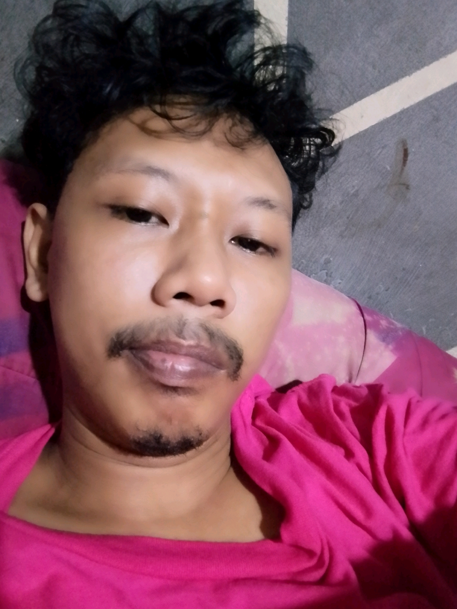 A close-up selfie of a man with dark, curly hair and a thin mustache and goatee is shown. He is wearing a bright pink t-shirt. The background appears to be a textured gray surface with a thin white line. The man's head is tilted slightly, and he is looking directly at the camera. The image seems to have been taken indoors, possibly in a bedroom, given the presence of what looks like a pillow with a pink and purple patterned fabric beneath his head and to his left. The overall lighting is somewhat dim.