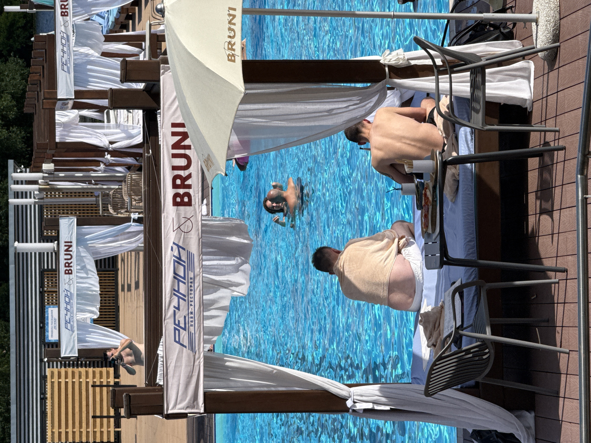 An overhead shot shows a man holding a phone to his ear and looking at it in the background. In the foreground, two men sit on lounge chairs by a pool, facing away from the camera. The man on the left is wearing a tan towel around his shoulders and white shorts. The man on the right is wearing black shorts. A table with food and drinks sits between them. White curtains hang from a wooden structure overhead, and a white umbrella is visible to the right. The pool is bright blue. A banner above the lounge area reads "РЕЧНОЙ BRUNI." This is a scene at the "RECHNOY" beach club in Khimki, Russia.