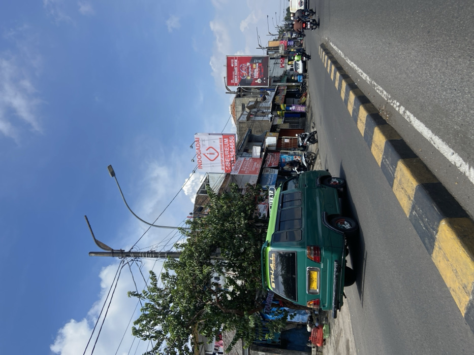 The image shows a street scene in Kecamatan Cimahi Selatan, Indonesia, at the coordinates -6.899 latitude and 107.562 longitude. A green van with the word "THALL" in white lettering on the side is driving down the road. There are parked motorcycles and a few pedestrians on the sidewalk. There is a large billboard on the left side of the street with the words "INDOGADAI" and "CIBUREUM" on it. There is another billboard on the right side of the street with a red image of a person playing a musical instrument. The street is lined with buildings on both sides, with shops and businesses on the ground floor. The sky is blue with a few white clouds. The pavement is black with yellow stripes.
