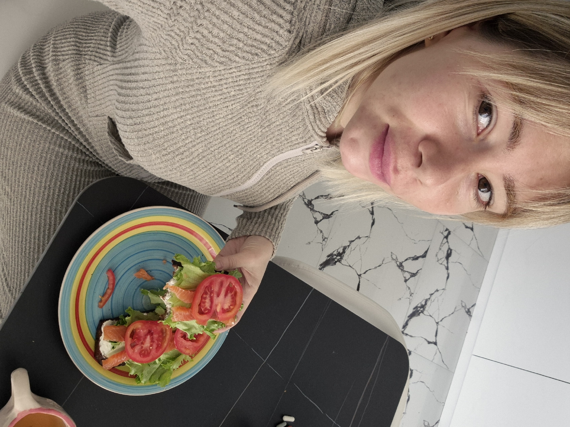 A fair-skinned blonde woman is holding a colorful plate with a sandwich. The sandwich is composed of bread, a white creamy spread, lettuce, slices of salmon, and slices of tomato. The plate has a colorful rim and is sitting on a black tabletop. A mug is partially visible on the bottom left. The woman is wearing a beige sweater. The location appears to be an interior space, possibly a kitchen in a residential area. The flooring is marble-like and a white cabinet is in the background. It is impossible to determine if the location is in Batumi, Georgia based on the information provided in the image.