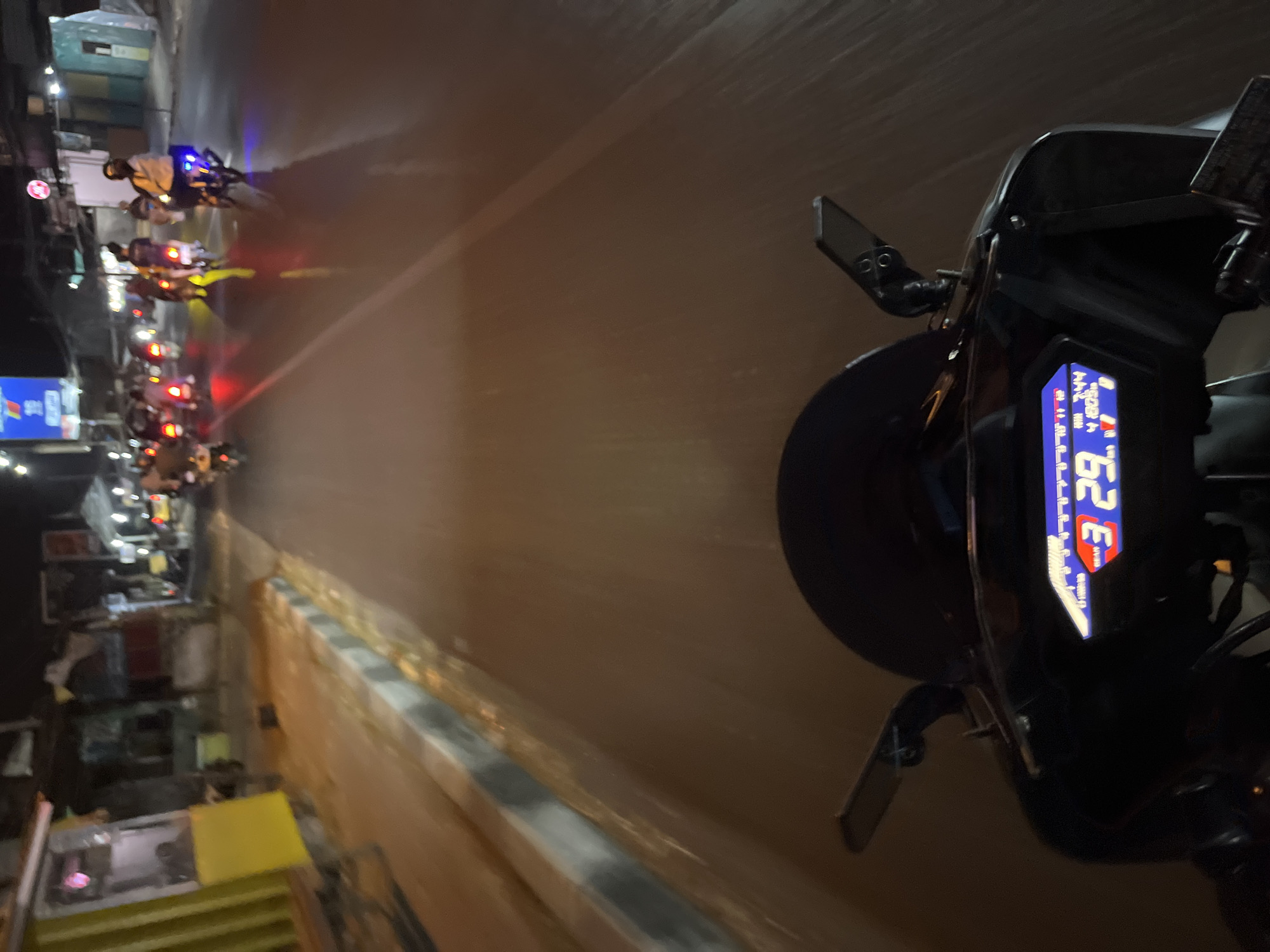 The image is taken from the perspective of a person riding a motorcycle at night in Kalinyamatan, Indonesia. The motorcycle's digital dashboard is visible in the foreground, displaying "3 29" and "4 1803". The road ahead is illuminated by streetlights and the headlights of other vehicles. Several motorcycles and their riders are visible on the road, some with blue and red tail lights. The surroundings include buildings and street vendors on the left side of the road. The overall scene depicts a typical street view at night in a bustling urban environment.