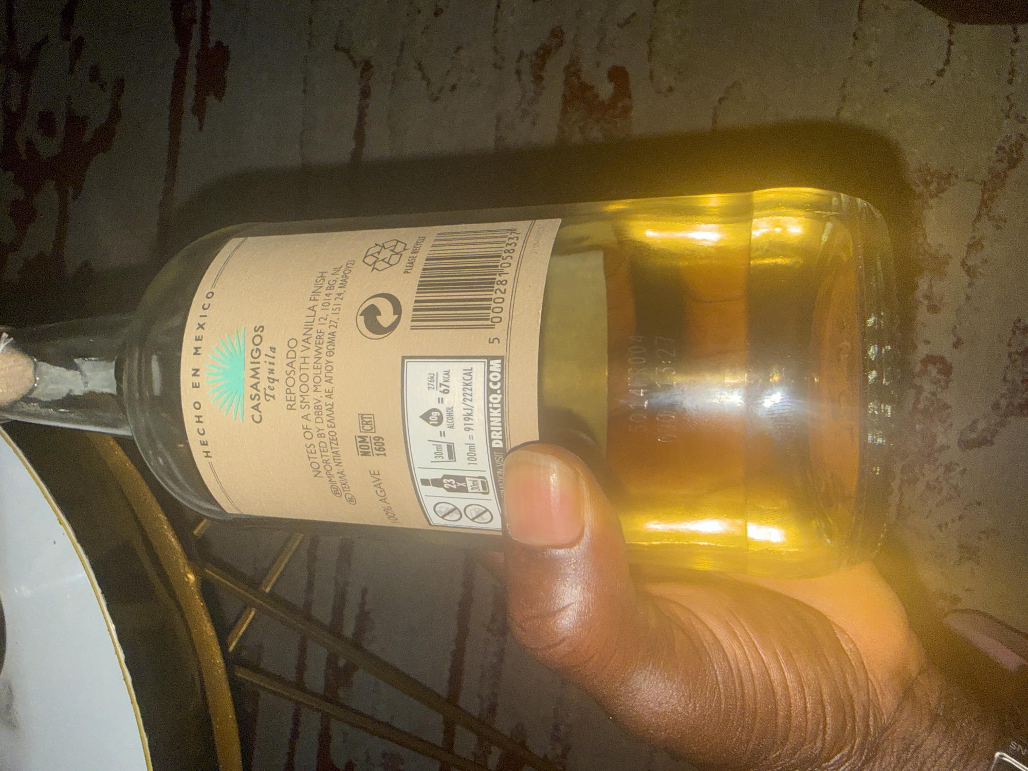 A hand is holding a bottle of Casamigos Tequila Reposado. The bottle is positioned horizontally with its label facing the viewer. The label indicates the tequila is "Hecho en Mexico" (made in Mexico) and contains notes of smooth vanilla finish. Nutritional information is printed on the label. The bottle also has a barcode. The surrounding environment appears to be indoors on what is possible a marble table. Location information (Igboefon, Nigeria) is not discernible from the image.
