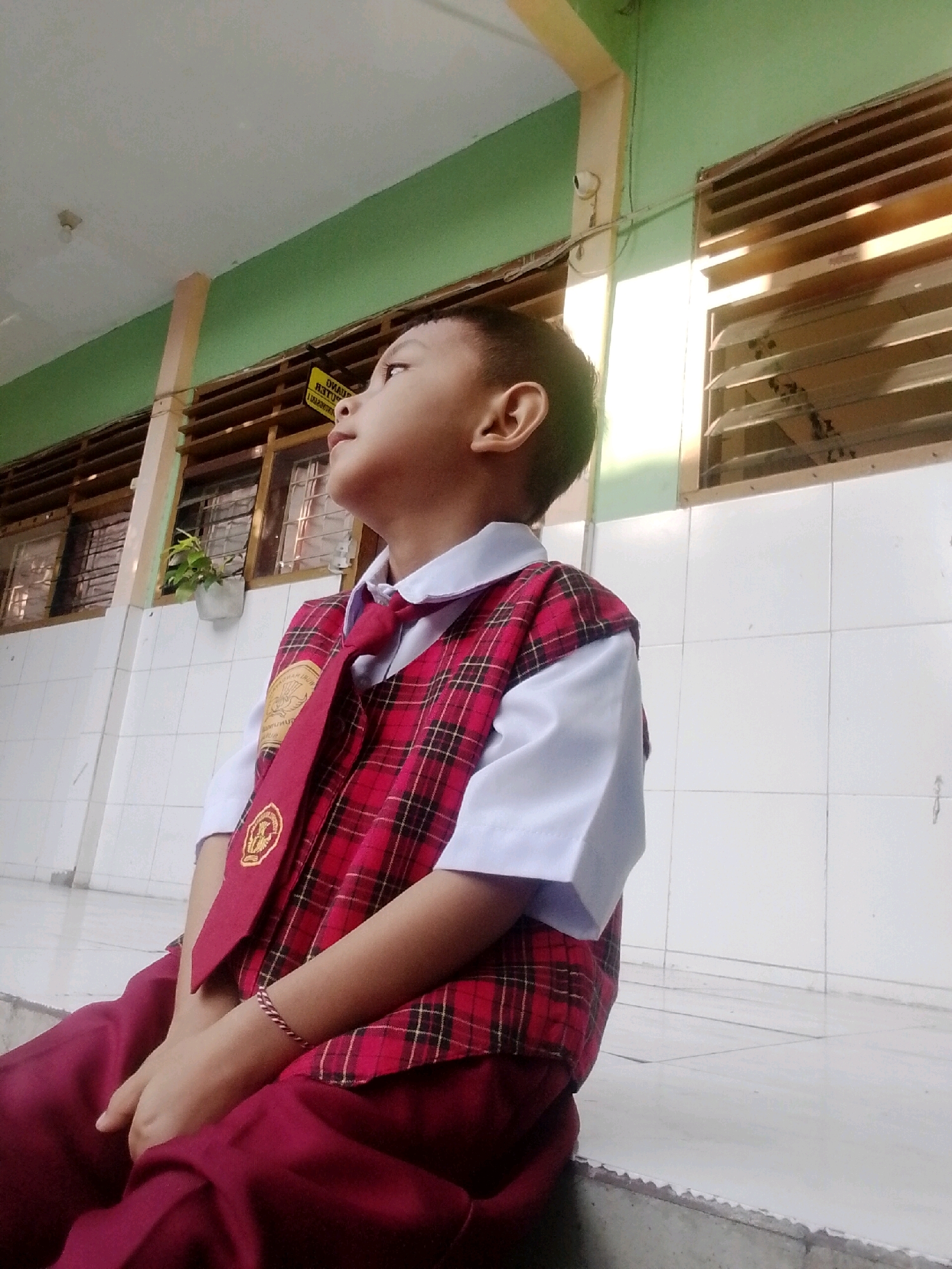 In the city of Surabaya, Indonesia, a young Indonesian boy, appearing to be of primary school age, is captured in a medium shot, seated and looking upwards. He is wearing a school uniform consisting of a white collared shirt, a red and black plaid vest, and a maroon tie with a school emblem. His maroon pants are visible, along with a thin, braided bracelet on his left wrist.
The background depicts an outdoor corridor or walkway of a school building. The walls are tiled in white squares, and a green-painted section runs along the top of the wall. Louvered windows with wooden frames are set into the wall, with sunlight filtering through. A small potted plant sits on a ledge beneath one of the windows. The floor appears to be tiled or paved. The overall lighting suggests it is daytime, with the sun casting strong highlights and shadows. The boy's gaze is directed towards the upper right of the frame, suggesting he is looking at something or someone outside the captured scene.