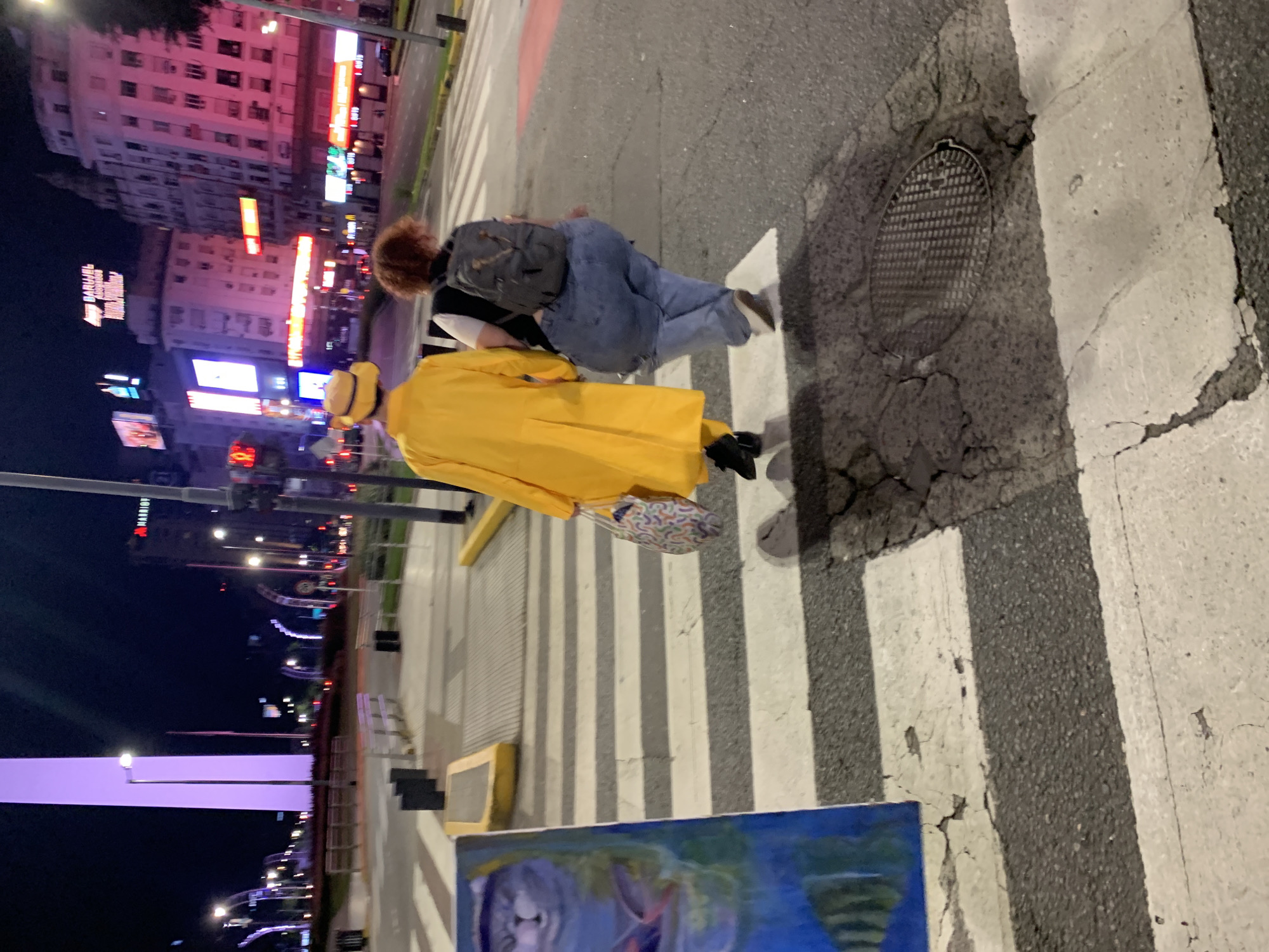 The photograph depicts two individuals crossing a street in Buenos Aires, Argentina. One person is wearing a bright yellow coat and hat and is carrying a patterned bag. The other person is wearing jeans and a backpack. They are walking on a crosswalk.
In the background, the Obelisco monument and buildings with illuminated signs are visible. There is also a painting or canvas on the sidewalk in the foreground. The street appears to be well-lit and the setting seems to be at night.