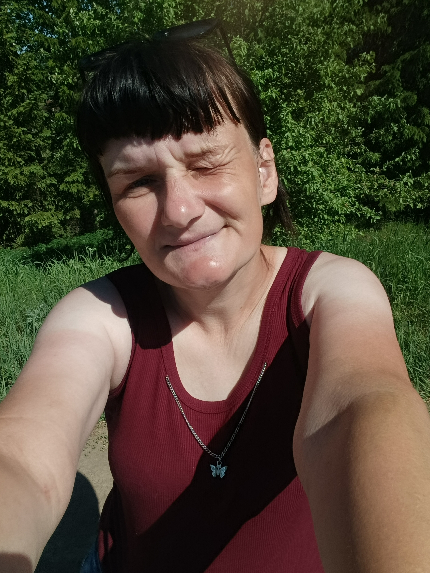 A woman is taking a selfie outdoors. She is wearing a maroon tank top and a silver chain with a butterfly pendant. Her hair is dark and has bangs, and she is wearing sunglasses on top of her head. She is winking one eye. The background consists of green trees and grass. The location appears to be in a park or natural area, city of Unknown, Unknown.