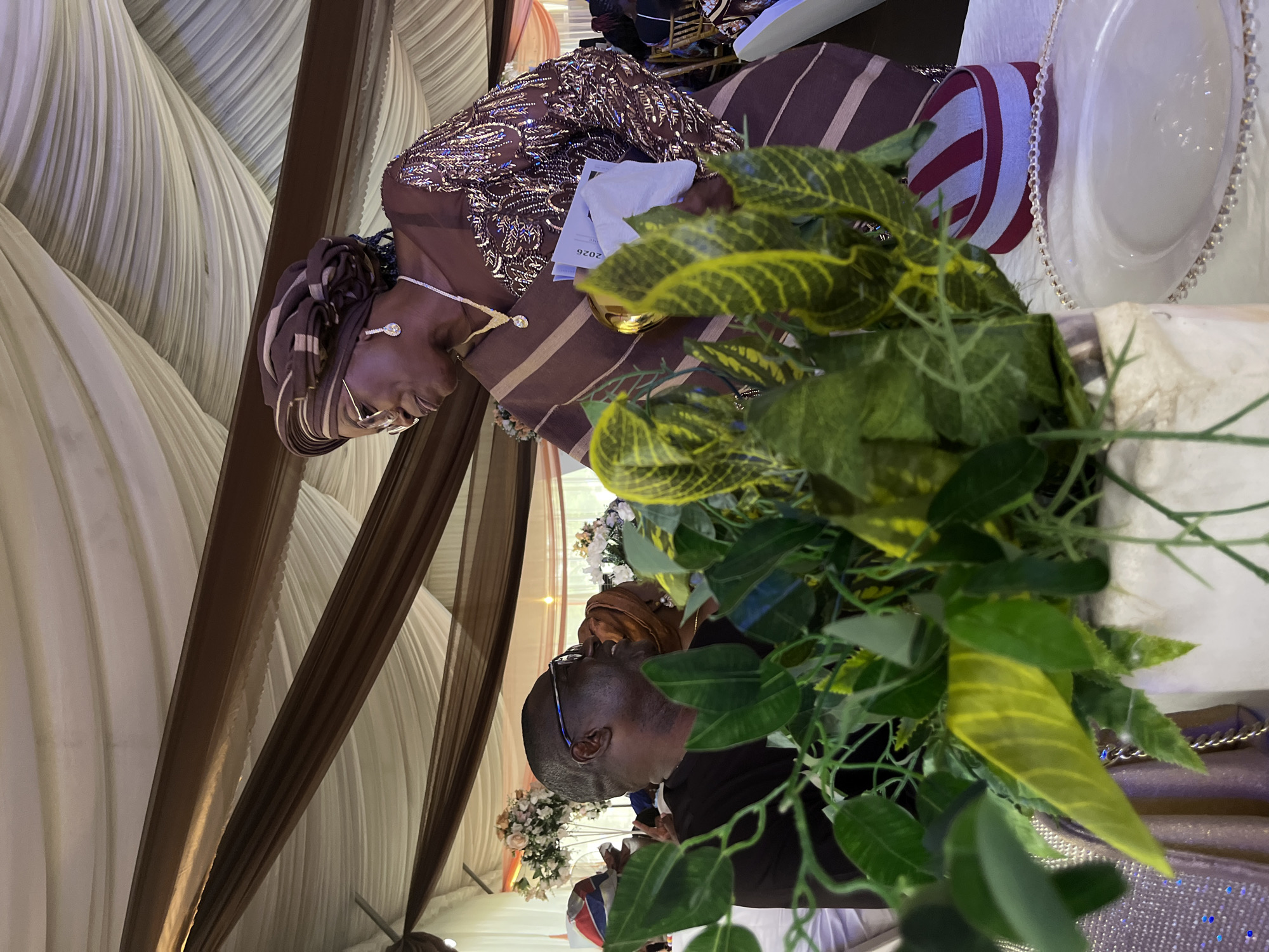 The image captures an indoor event, likely a celebration or reception, in Abuja, Nigeria, featuring elaborate decor and attendees. The ceiling is dramatically draped with flowing white fabric, accented by rich brown panels.
In the foreground and mid-ground, two individuals are seated at a table, partially obscured by a vibrant green and yellow foliage centerpiece. A woman, prominently featured, wears a meticulously embellished brown gown adorned with gold sequins and intricate patterns. She complements her attire with a matching brown and striped headwrap (gele), a sparkling silver necklace with a large pendant, and round earrings. She appears to be looking down or to the side.
Behind the foliage and to the right, a man wearing glasses is partially visible, seated at the table. He is dressed in a dark top.
The table setting includes white plates and a decorative beaded chain. A distinct red and white striped cylindrical object is also on the table. A white card is placed near the woman's arm, on which the number "2026" is clearly visible. In the background, elegant floral arrangements with white and light pink flowers can be seen. The lighting suggests an indoor evening event, contributing to a celebratory atmosphere.