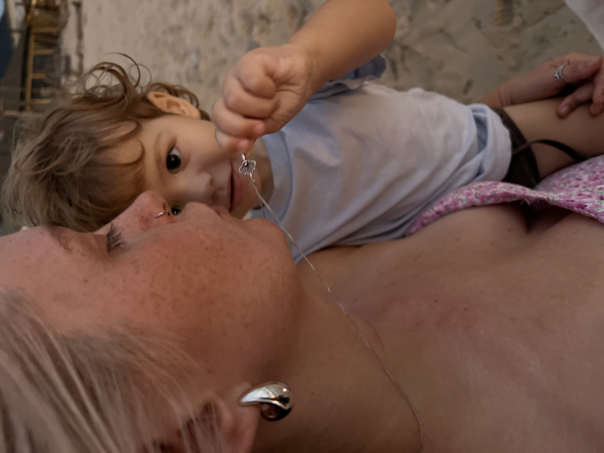 In this close-up, intimate shot, a young child leans over an adult, their faces nearly touching. The child, with curly brown hair and wide, dark eyes, is focused on a delicate silver necklace with a butterfly pendant that they are holding. The adult, with blonde hair and freckled skin, gazes upwards, their expression soft and engaged. The child is wearing a light blue t-shirt, while the adult is wearing a pink and white floral patterned garment. A silver earring is visible on the adult's ear. The background is blurred and appears to be a textured surface, possibly a couch or floor. The lighting suggests it is daytime, and the overall mood is one of tenderness and connection.
