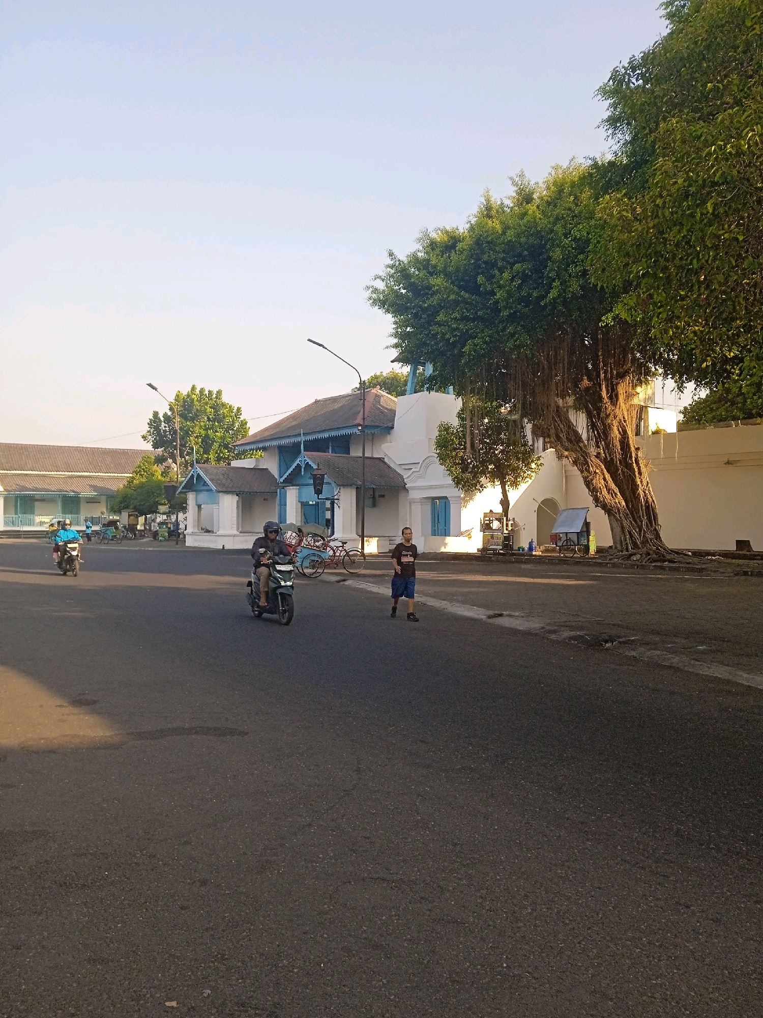 In Surakarta, Indonesia, a street scene unfolds with a prominent banyan tree casting dappled sunlight on an asphalt road. A person jogs on the sidewalk, while two individuals on motorbikes navigate the street. In the background, traditional Indonesian buildings with white walls and blue trim line the road, suggesting a historic or culturally significant area. The sky is clear and bright, indicating daytime.