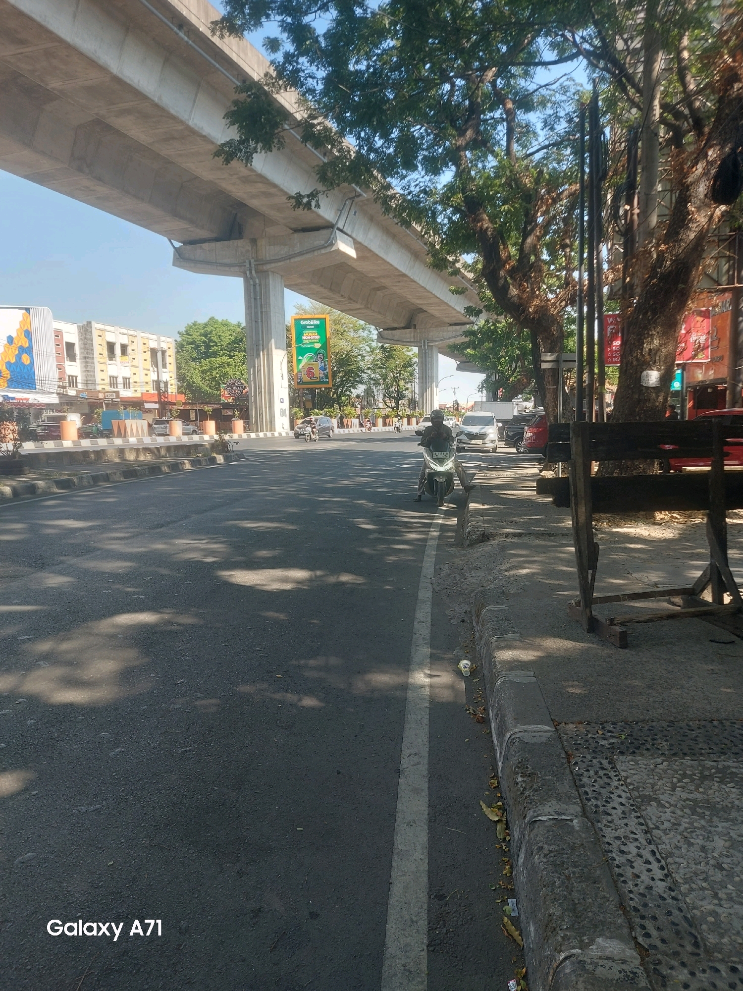 In Makassar, Indonesia, a motorcycle rider wearing a helmet is seen traveling on a paved road under a concrete overpass. The road features a white dividing line. To the right of the road is a sidewalk with a stone pattern and a wooden barrier. Several trees line the sidewalk, their leaves casting shadows on the ground. In the background, buildings and a billboard are visible. The sky is clear and blue.