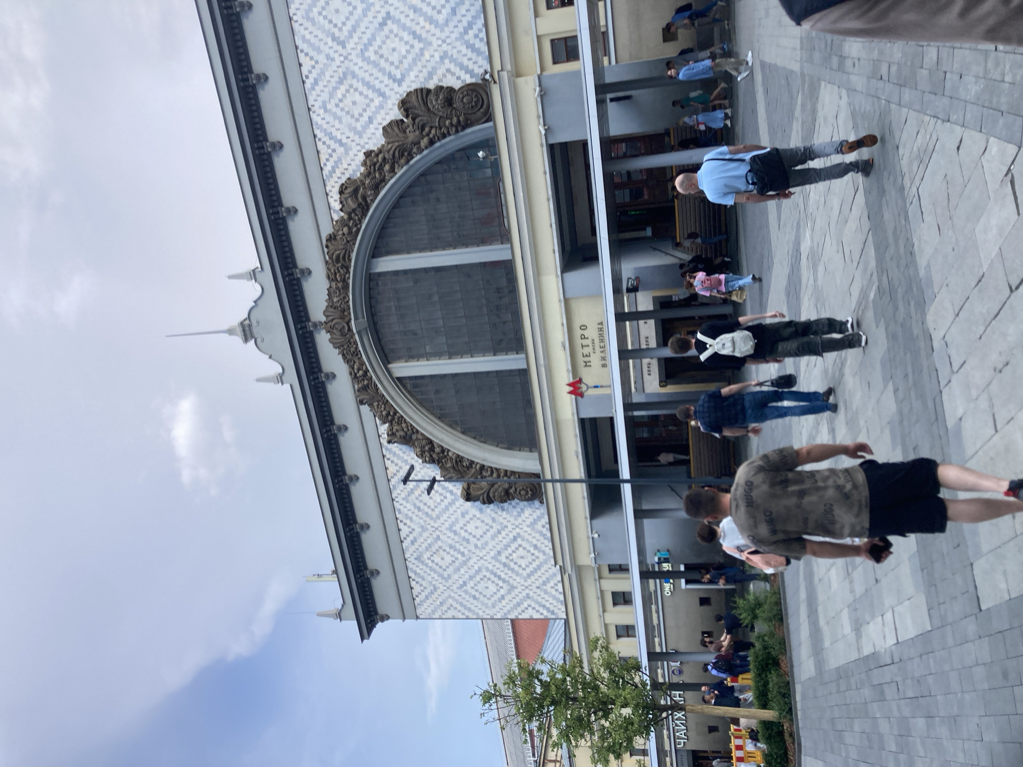 The image shows a street scene in Moscow, Russia, with people walking on a paved plaza in front of a building with ornate architecture. The building features a large arched window with decorative stonework and a patterned facade. Several people are visible, some walking towards the viewer, others away. One person in the foreground is wearing a camouflage t-shirt and black shorts. Another person in a light blue shirt and grey pants is walking to the right. The scene appears to be during the daytime, with a bright sky visible. The paving stones of the plaza create a geometric pattern. Text is visible on the building, including "МЕТРО" and "В. И. ЛЕНИНА", indicating it is a metro station. Other partial text on signs is also present but not fully legible. The overall atmosphere seems to be that of a busy public space.