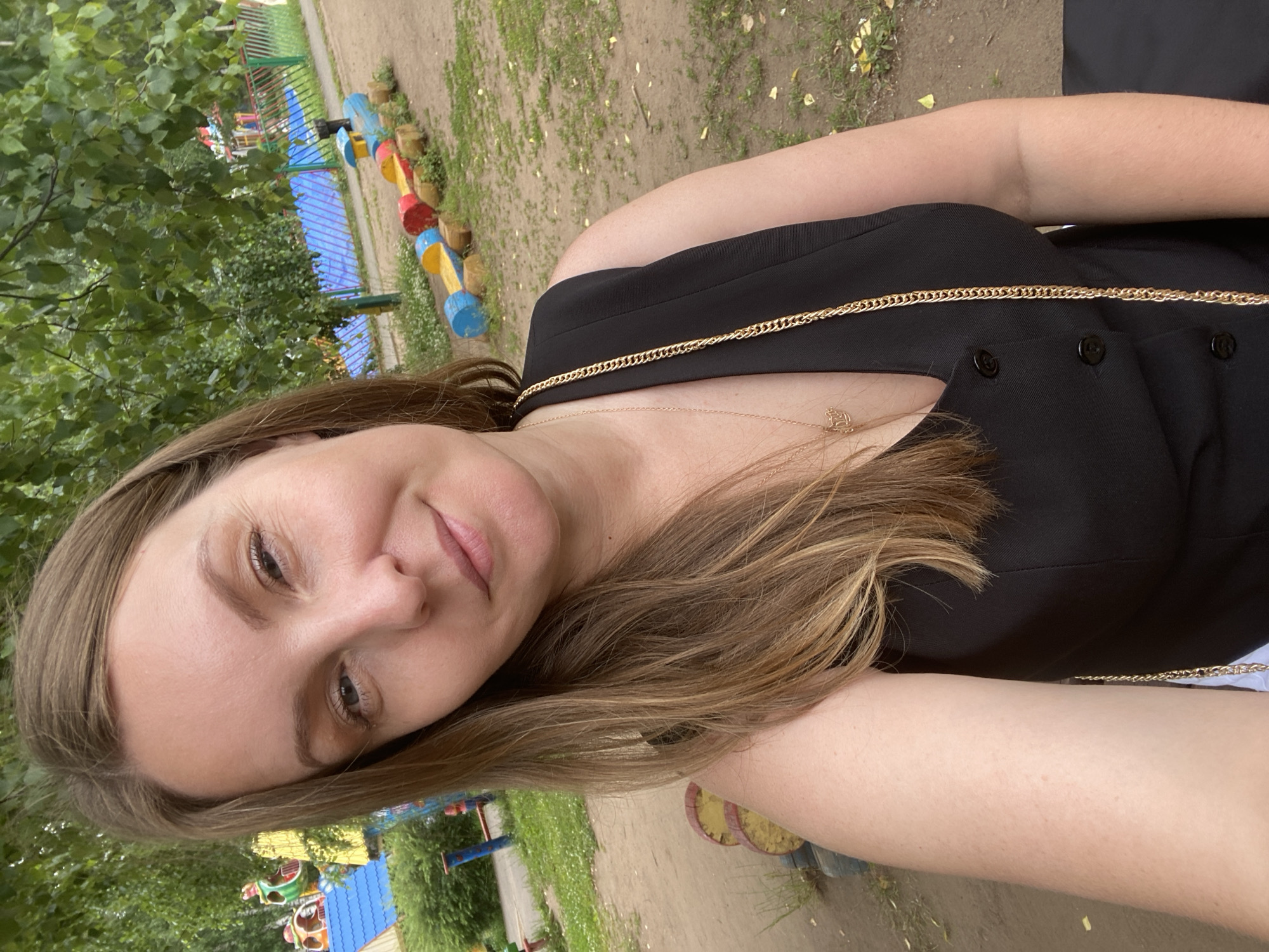 A woman with shoulder-length brown hair takes a selfie outdoors. She is wearing a black sleeveless vest with buttons and a gold chain necklace with a pendant. Behind her, a playground is visible with colorful equipment, including a wooden train structure and blue and yellow elements. Trees with green leaves form the background. The ground is sandy and patchy with grass.