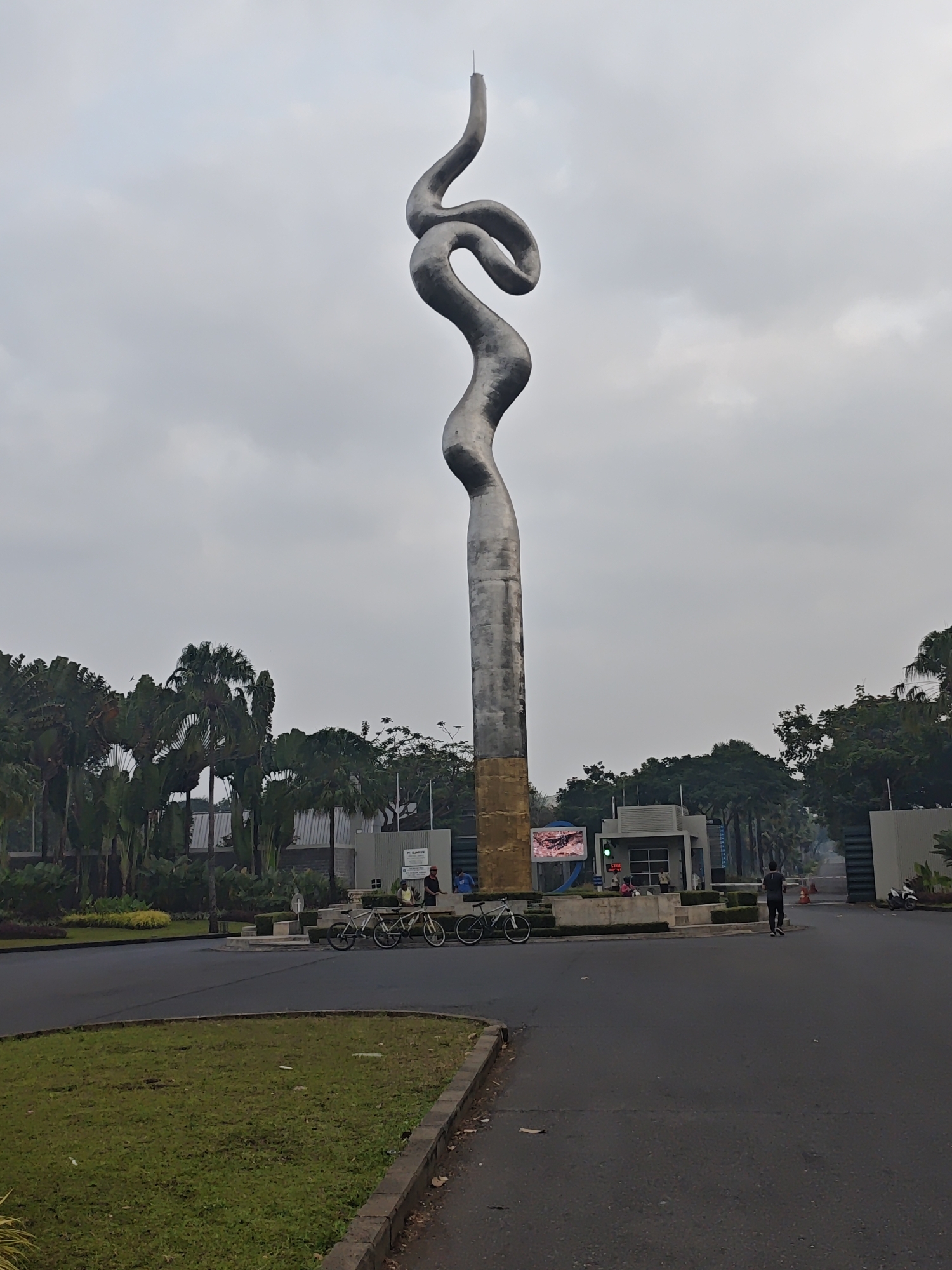 This is a full-shot, eye-level, outdoor photograph taken on an overcast day in the city of Bacin, Indonesia. The primary subject is a large, abstract, metallic sculpture that resembles a twisted ribbon or a stylized flame. The sculpture is silver and stands on a golden-colored cylindrical base. It is positioned in the center of a paved roadway, in front of a modern building complex with a security gate.
To the left of the sculpture, there is a well-maintained green lawn bordered by a paved walkway. Behind the lawn and to the left of the sculpture, a cluster of lush green palm trees and other tropical foliage is visible.
In the mid-ground, to the left of the sculpture, three bicycles are parked side-by-side. Near the building complex, several people are present. A few individuals are seated on a low wall in front of the building. One person is standing and appears to be running or jogging on the roadway to the right of the building. Another person in a black shirt and dark pants is walking away from the camera towards the right side of the frame.
The building complex features a modern architectural style with clean lines, gray walls, and glass elements. A digital billboard is mounted on the left side of the entrance, displaying a colorful image. A traffic light is visible above the billboard, showing a green light. A sign reading "STOP" is also present.
In the background, more trees and foliage are visible, and the sky is heavily overcast with gray clouds. The overall impression is a contemporary urban environment with a prominent piece of public art.