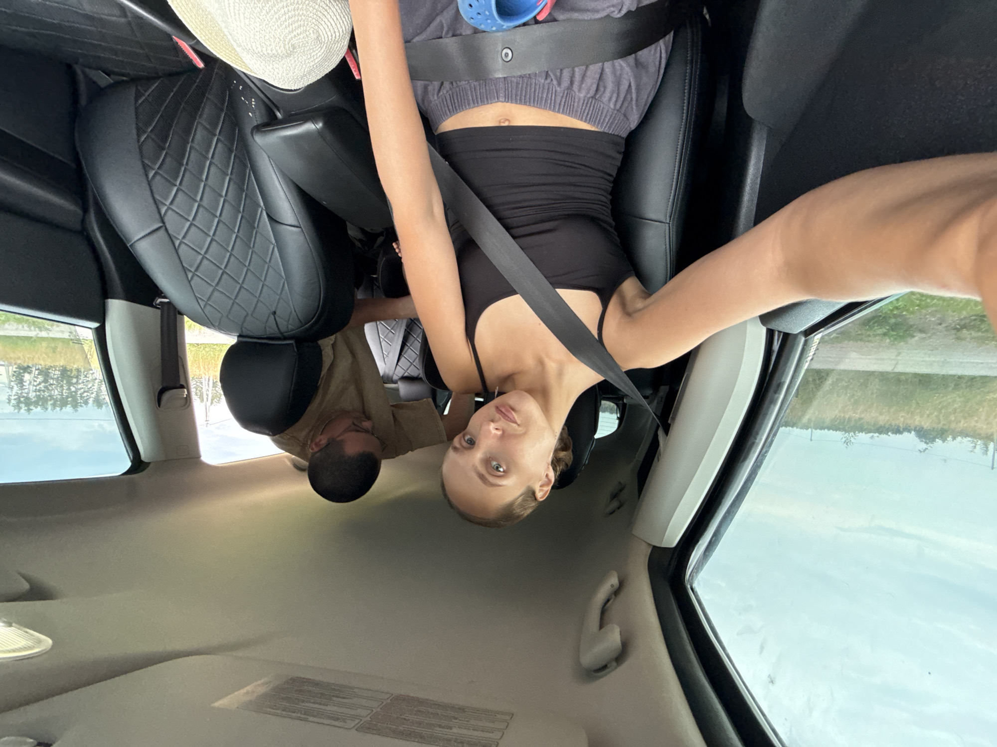 Two individuals are inside a vehicle. A woman with light brown hair pulled back is in the driver's seat, wearing a black tank top and grey sweatpants. She is wearing a seatbelt and her left arm is extended towards the camera. A man with short dark hair and glasses is in the passenger seat, wearing a light brown t-shirt. He is looking down and appears to be reaching for something. The interior of the car is visible, including black leather seats with a diamond pattern on the passenger seat. Outside the car, a grassy field and trees are visible under a cloudy sky. The location is not specified in the image, and therefore cannot be confirmed as the city of Подшивалово, Russia.
