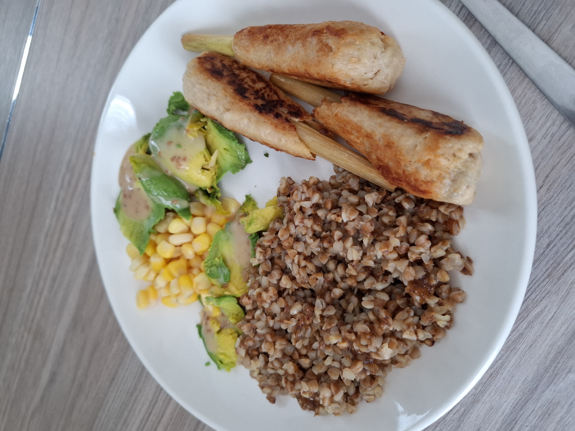 A white plate of food sits on a grey wooden surface. The plate contains a portion of cooked buckwheat, a salad of sliced avocado and corn with a light-colored dressing, and three cylindrical food items that appear to be meat or vegetable patties, each skewered on a wooden stick. The patties are lightly browned with some char marks. The background is blurred, but hints of a reflective surface and a light-colored wall are visible. No people or specific city landmarks are identifiable.