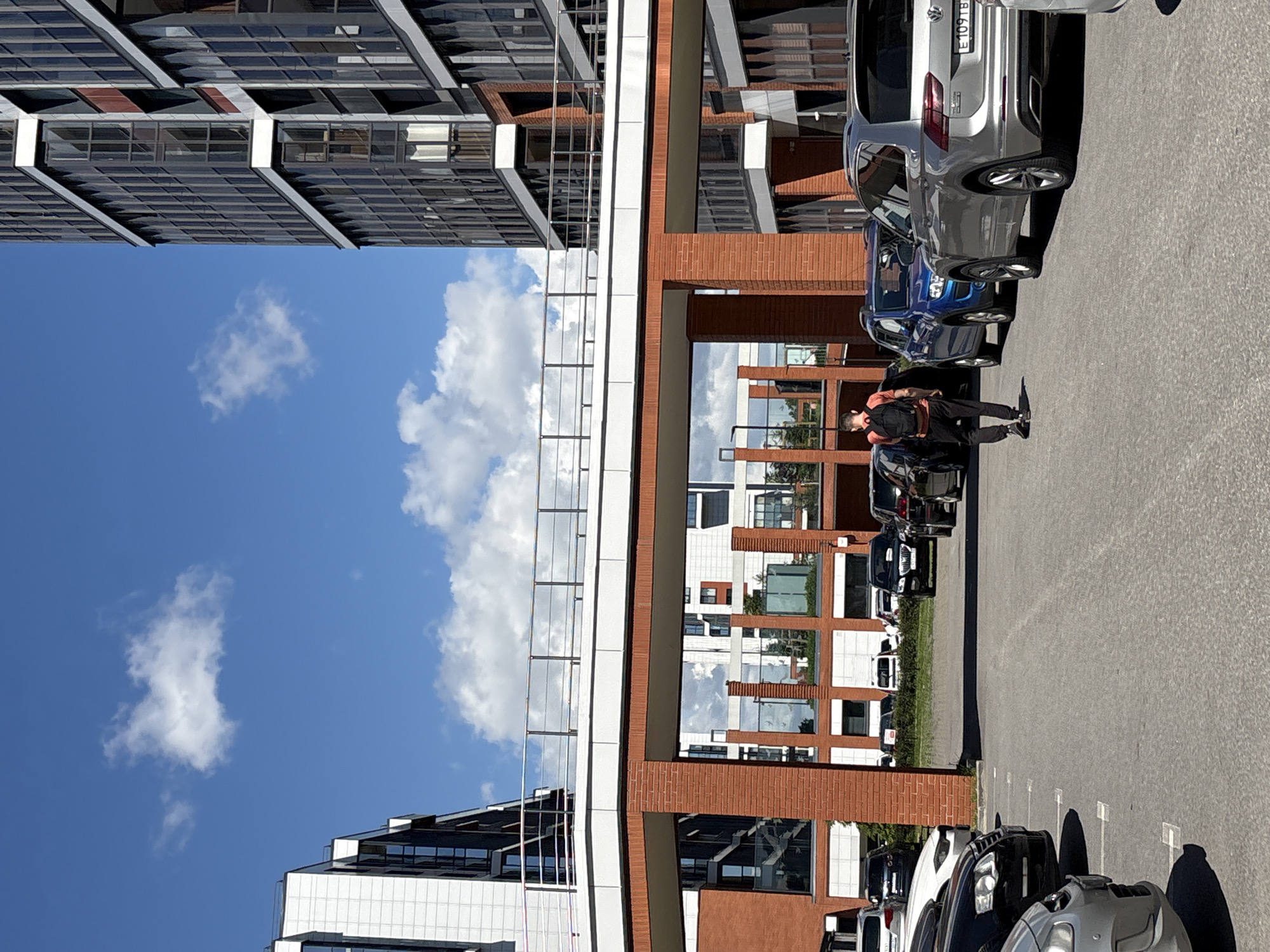 The image captures a street scene in Saint Petersburg, Russia, featuring a modern building with a glass and brick facade. Several cars are parked in a lot alongside the building, with visible parking spaces marked on the asphalt. A person, seen from behind, is walking away from the camera, carrying a backpack. The sky is blue with scattered white clouds, suggesting fair weather during the daytime. The building's reflective windows mirror the sky and surrounding structures, adding depth to the scene. The architecture of the building appears contemporary, with clean lines and geometric shapes. The overall impression is one of an urban environment bathed in natural light, showcasing a typical day with people and vehicles interacting within the architectural landscape.