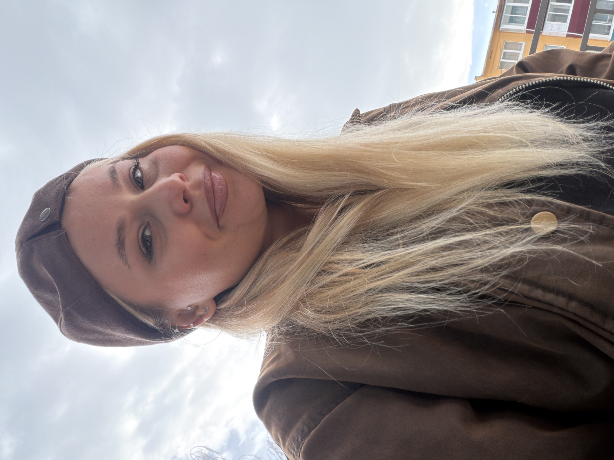 A female individual is depicted in a close-up, upward-angle portrait, likely a selfie, against an overcast sky. The subject has long, straight blonde hair and fair skin. She is wearing a brown beret with a metallic button-like embellishment on the left side of her head. Her visible left ear features multiple piercings, including several small stud earrings on the helix, conch, and lobe areas. She is wearing a brown collared jacket, partially unzipped, over a dark-colored top. The individual is looking directly at the camera with a subtle smile. In the lower right background, a multi-story building is partially visible, characterized by yellow and burgundy/red sections with white-framed windows and balconies. The specific location within the city of Sarapul, Russia, cannot be definitively identified from the visual information presented in the image.