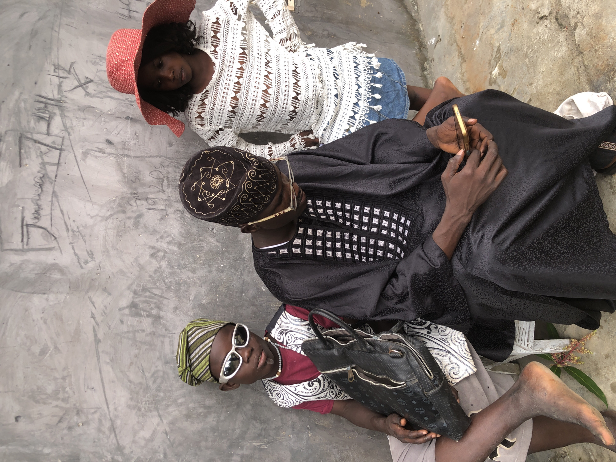 Three young people are posing against a weathered concrete wall with graffiti. A boy in the foreground on the left wears a green and white striped cap, large white sunglasses, a red shirt, and a patterned vest. He has a black bag on his lap and is barefoot. To his right, a person wearing a black cap with gold embroidery and glasses, dressed in a black traditional garment, looks down at a smartphone in their hands. To the right of the person in black, a girl wearing a red wide-brimmed hat and a white lace top over a denim skirt stands with her hands on her hips. The scene appears to be outdoors in an urban setting, possibly in Ado, Nigeria, indicated by the context provided. The wall behind them is textured and marked with visible graffiti.
