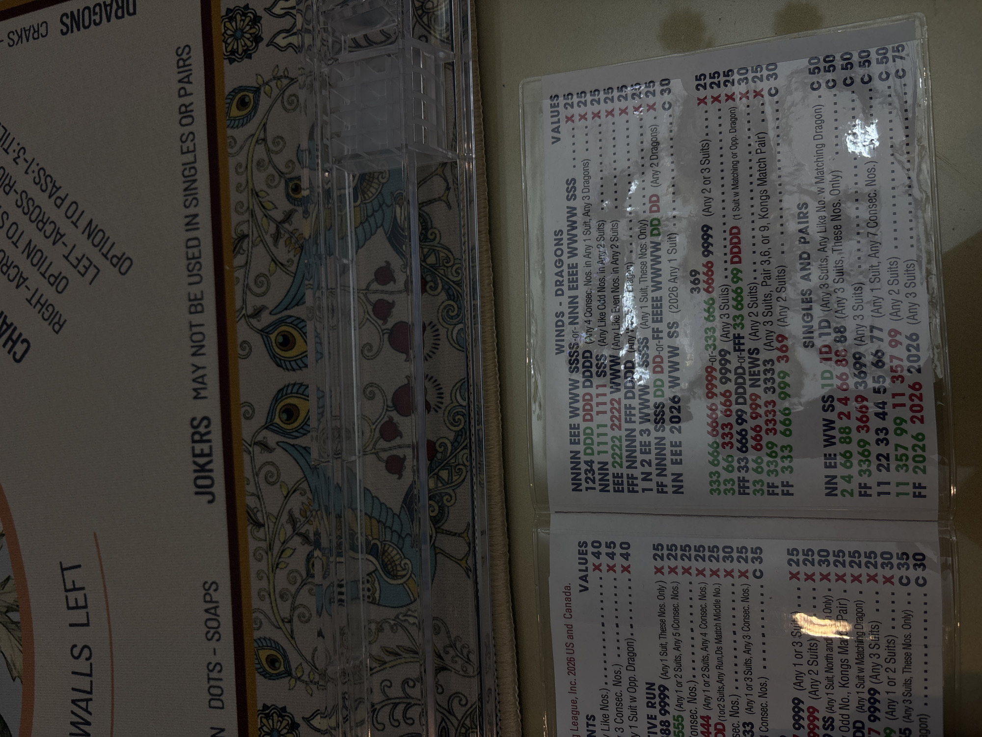 The image is a close-up shot of a mahjong set and a card detailing point values. The card is titled "Winds - Dragons" and "Singles and Pairs" and lists various tile combinations and their corresponding point values. The tiles themselves are not visible, but a plastic tile rack is partially visible in the upper center of the frame. The image is on a surface with a floral pattern. There is no indication of the location of Huixquilucan de Degollado, Mexico.