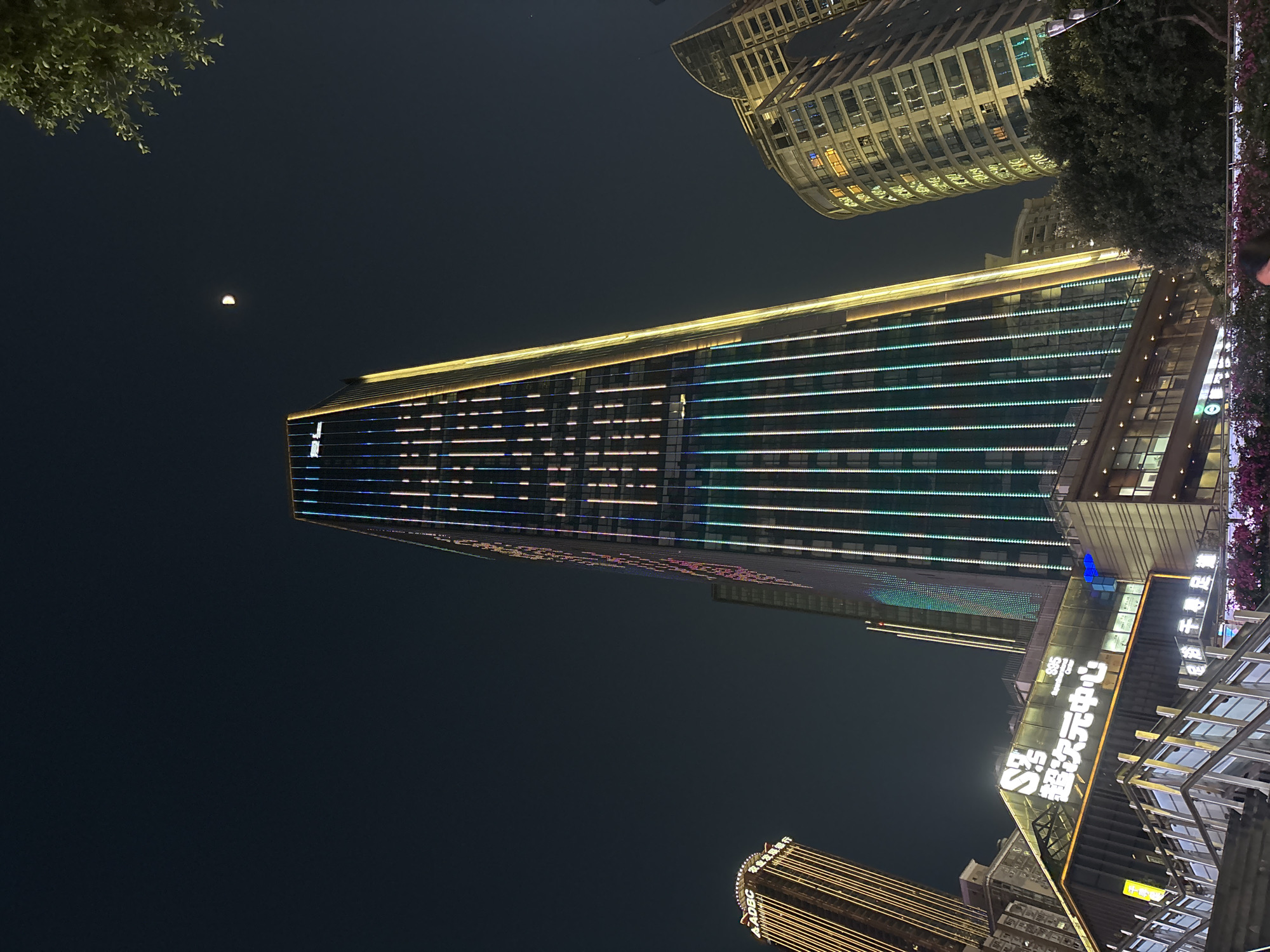 The image shows a nighttime urban scene in the Yuzhong District, China. A tall, modern building is the dominant subject, illuminated with horizontal rows of white, blue, and green lights. A smaller, curved building with illuminated windows is visible in the background. Street level, signage in Chinese characters is visible. The sky is dark, with a crescent moon visible in the upper left corner. The buildings and illumination indicate an urban environment.