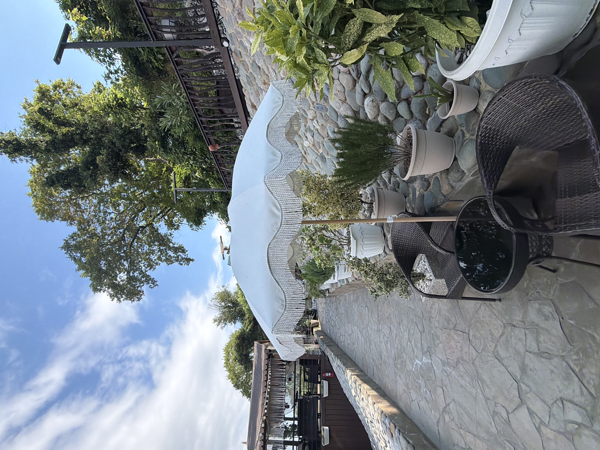 The image depicts an outdoor seating area at what appears to be a restaurant or cafe in Nizhnyaya Shilovka, Russia. A white fringed umbrella shades a stone-paved walkway lined with potted plants. Two wicker chairs and a small dark table are positioned near the walkway. In the background, a building with a wooden deck and thatched roof is visible. The area is situated on a hillside covered with lush green trees and a stone wall, suggesting a scenic location. The sky is blue with scattered white clouds. No people are present in the image.