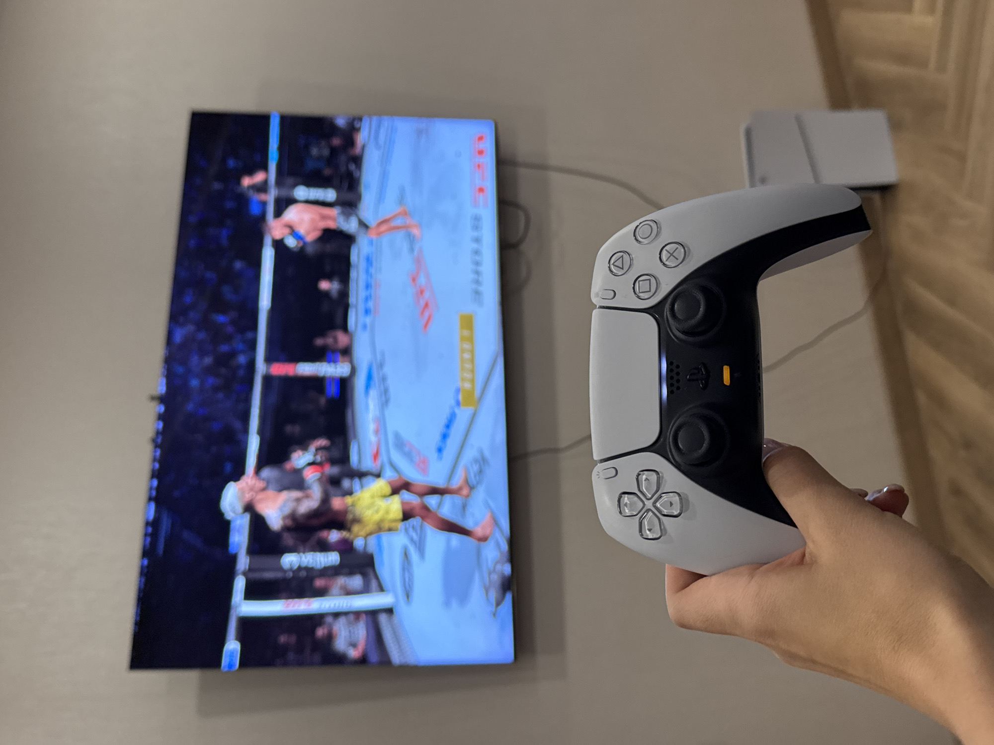 A person holds a white PlayStation Portal handheld gaming device in their hand. The screen of the device is active and displays a mixed martial arts (MMA) fight. A flat screen television is mounted on the wall behind the handheld device, also showing an MMA fight. A white charger for the device is positioned on the wooden surface next to the wall. The location is unknown.