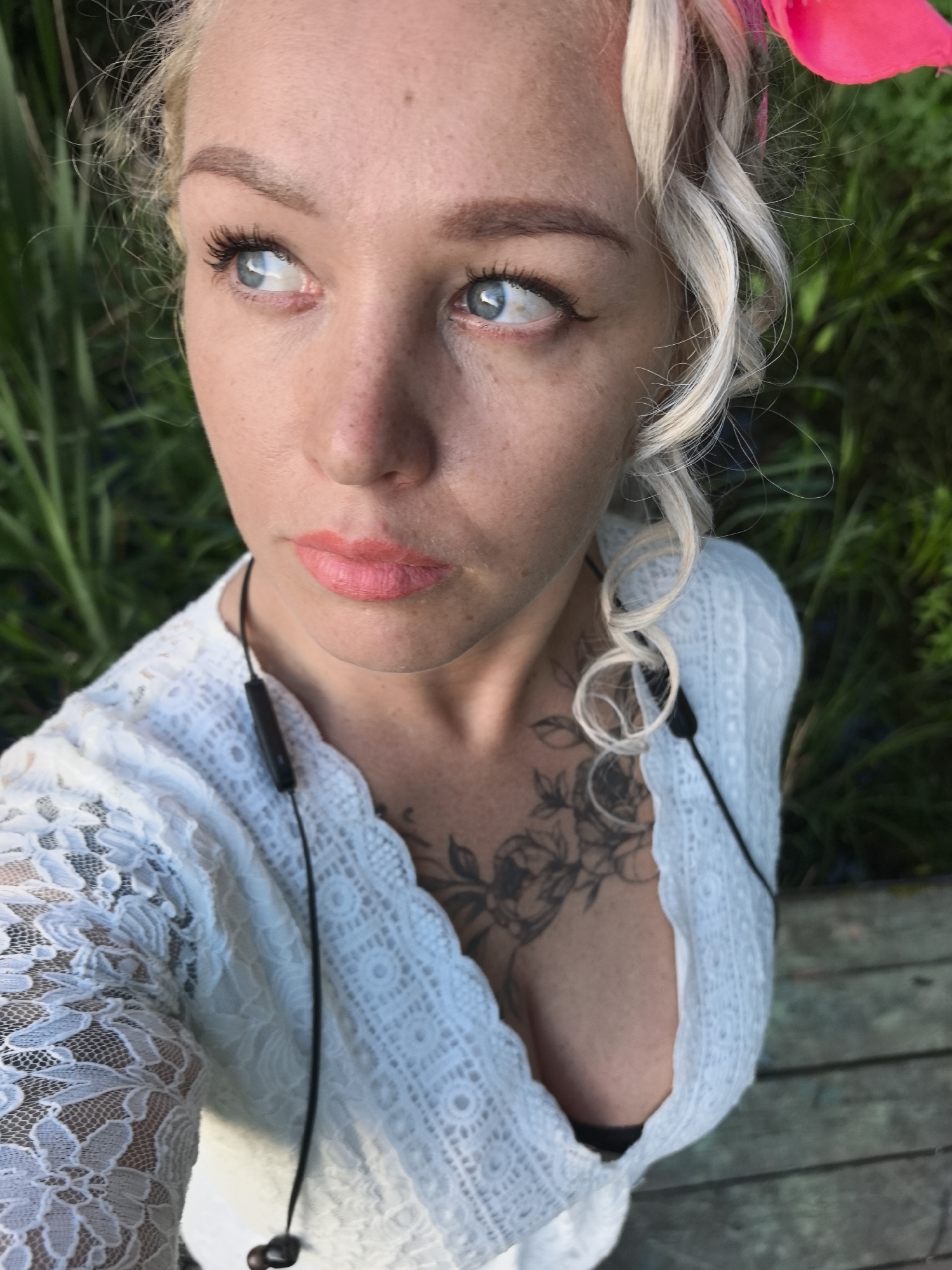 This is a high-angle, close-up selfie of a woman with blonde hair, adorned with a bright pink flower accessory. She is looking to her left, with a thoughtful or distant expression. The woman has light blue eyes, noticeable eyelashes, and subtle freckles on her skin. Her lips are painted with a coral-pink lipstick. She is wearing a white, lace-trimmed garment with a deep V-neck, revealing a tattoo of roses on her chest. Black earbuds are visible, with wires running down her chest. The background consists of lush green vegetation, suggesting an outdoor setting. The lighting indicates it is daytime, likely sunny, given the bright illumination. The wooden planks visible at the bottom right of the image hint at a pier or a deck. The overall mood is serene and natural. There is no visible text.