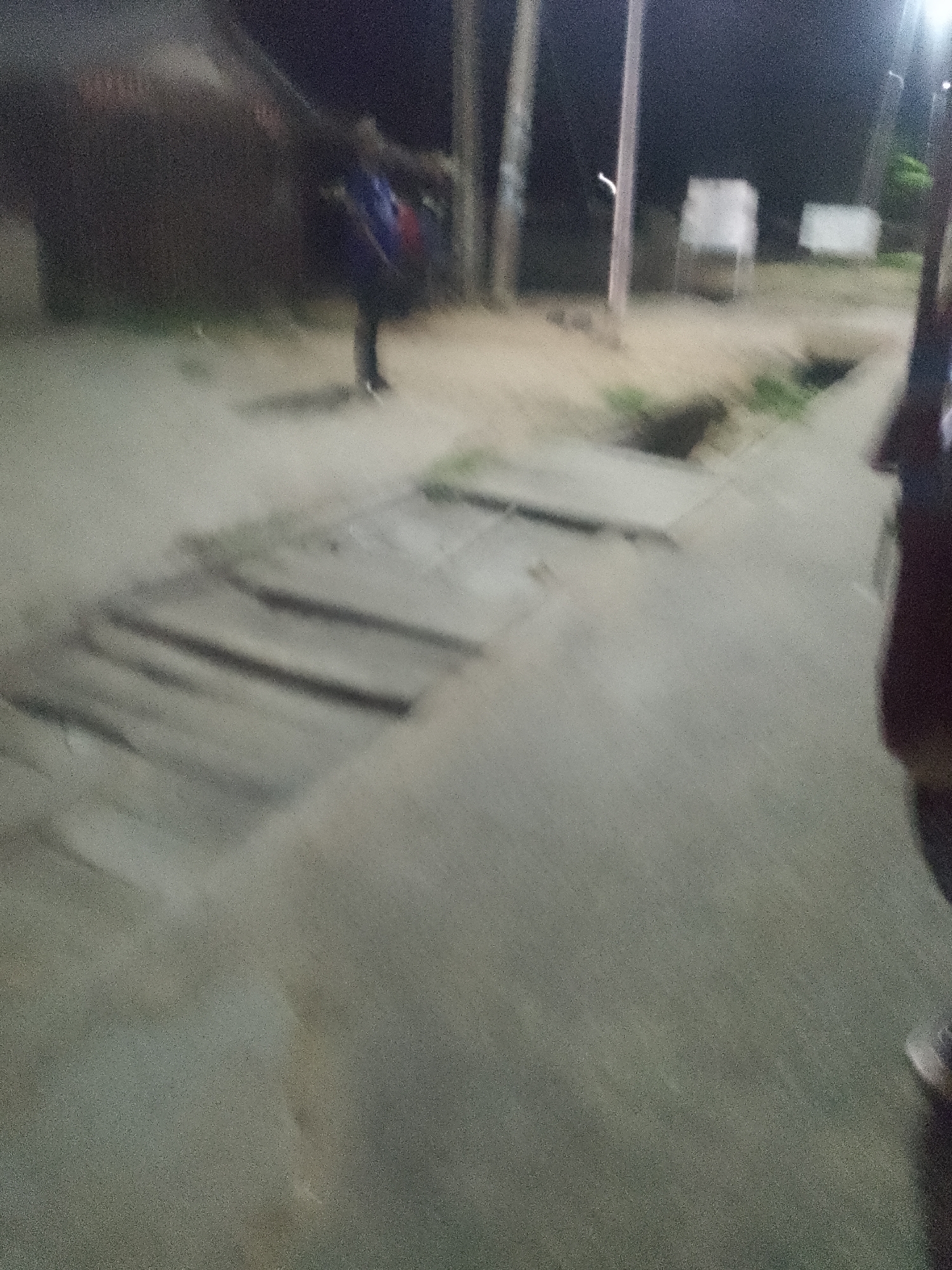 The image depicts a dimly lit outdoor scene, likely at night, with a sandy or dirt ground. In the background, there is a person standing, wearing a backpack. The person appears to be walking away from the camera. There are some blurry structures in the background, including what looks like a building and utility poles. The foreground shows a textured ground with what appears to be a manhole cover or drainage grate. The overall impression is that of a street or pathway in a somewhat undeveloped or rural area. The lighting suggests artificial illumination, possibly from streetlights or nearby buildings, casting long shadows and creating a grainy quality in the image. The environment seems to be within the city of Jalingo, Nigeria, given the context provided.