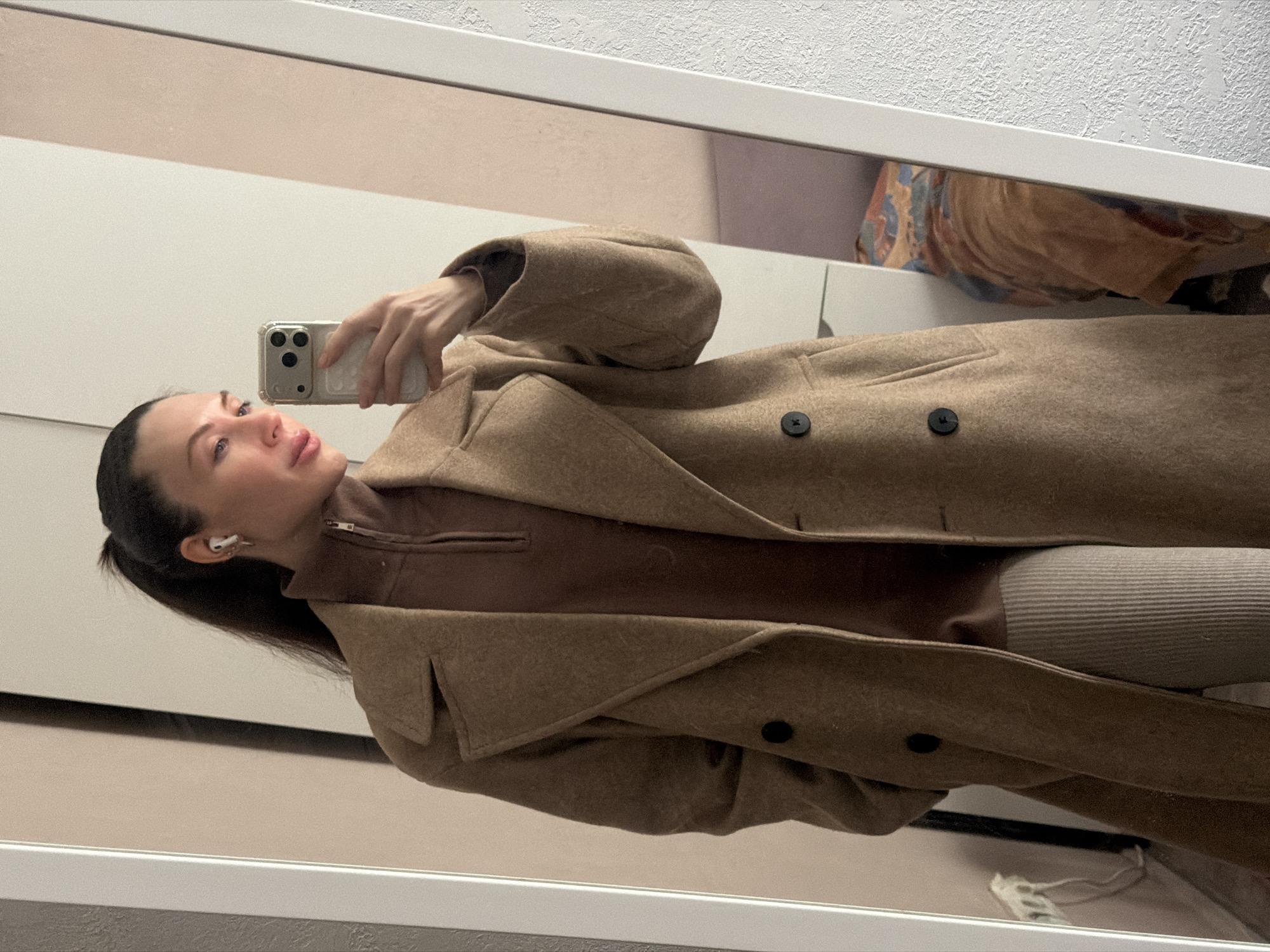 A woman is taking a mirror selfie indoors. She is wearing a light brown coat over a brown zip-up sweater and light grey ribbed pants. She has dark hair pulled back in a ponytail and is wearing an earbud in her left ear. She is holding a cell phone in her right hand. The background shows a glimpse of a room with white walls and a hint of patterned fabric. The location is purportedly in the city of Perm, Russia.