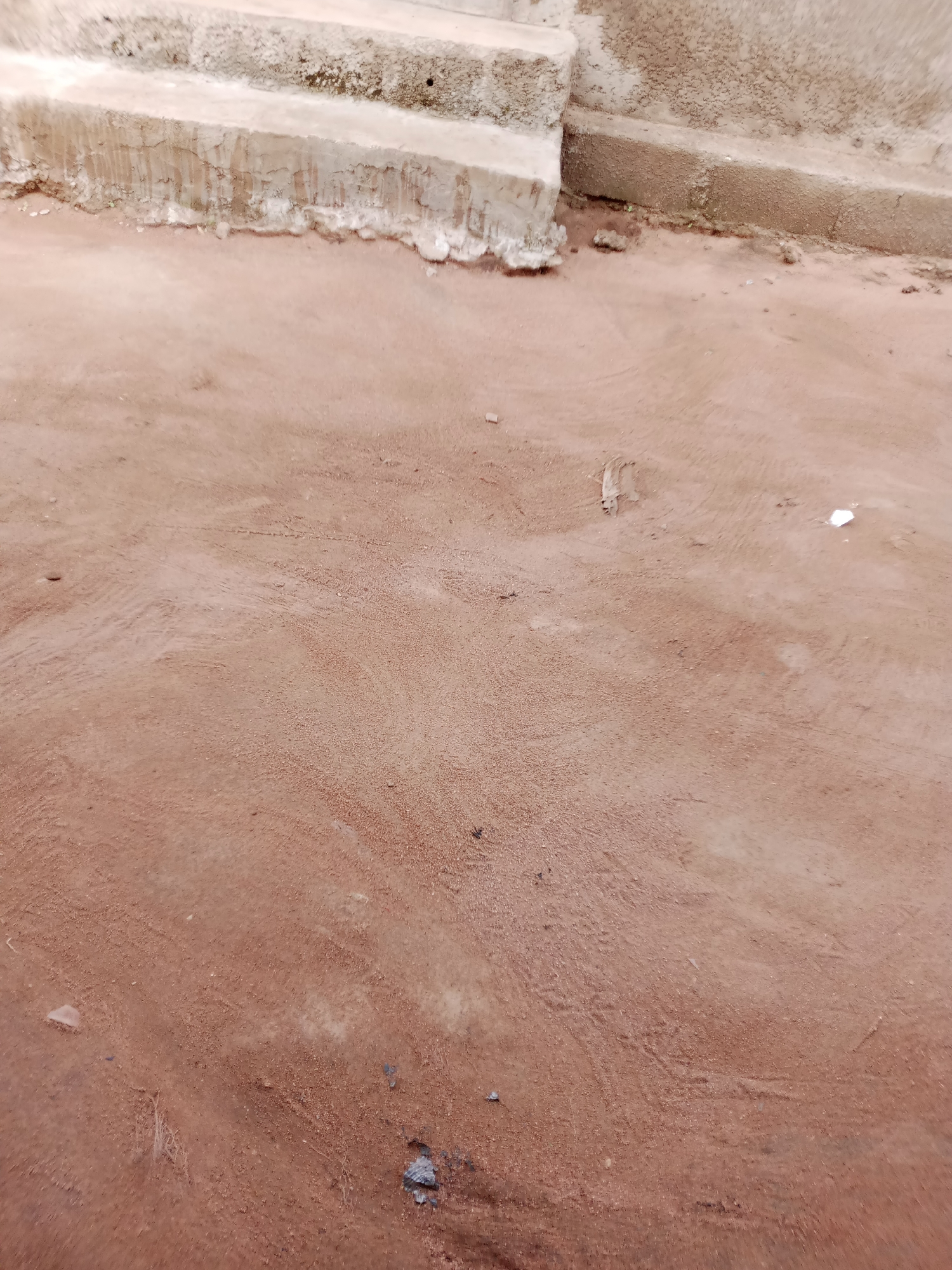 The image depicts a close-up view of a dirt ground with some concrete steps in the upper left portion of the frame. The ground is a reddish-brown color and appears to be composed of sand and fine gravel. There are some subtle textures and tire tracks visible on the ground. Small debris, such as small rocks and a piece of white plastic, are scattered across the surface. The concrete steps show signs of wear and tear, with chipped edges and some crumbling texture. The environment appears to be an outdoor setting, likely in a somewhat undeveloped area.