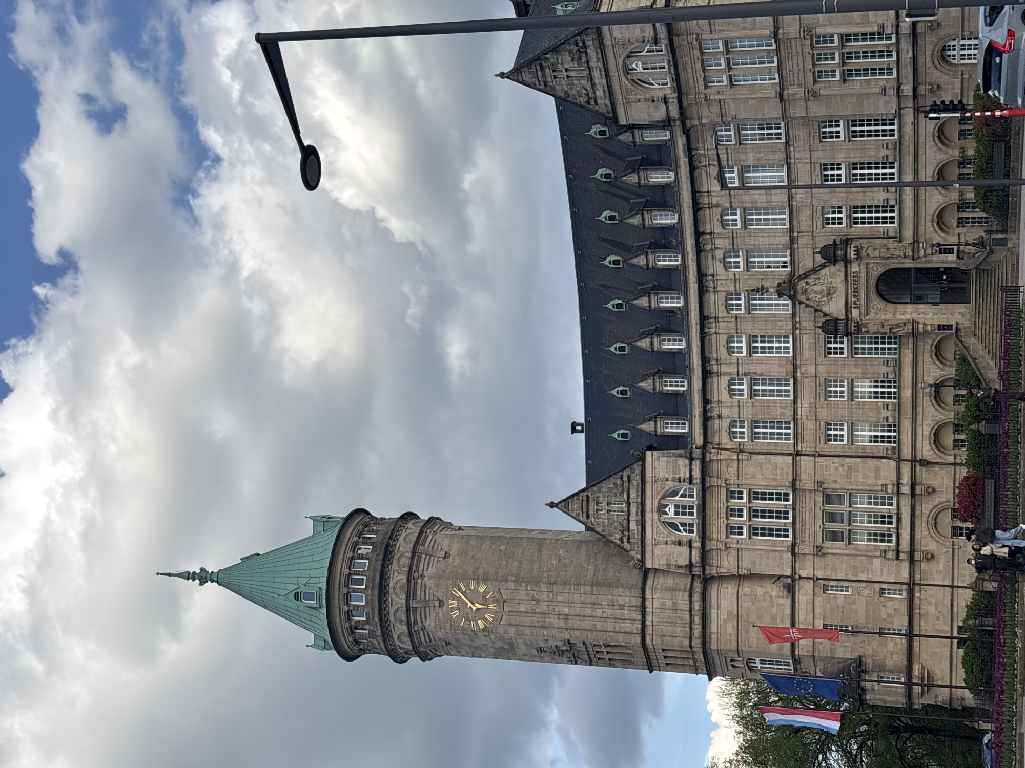 This image shows a low-angle view of the Grand Ducal Palace, located in Luxembourg City, Luxembourg. The architectural style is Flemish Renaissance. The tower on the left side displays a clock. Flags representing Luxembourg, the EU, and other entities are visible in the foreground. A lamppost and cloudy sky are visible above. Some pedestrians are on the right side of the building. A car can be seen in the upper right.