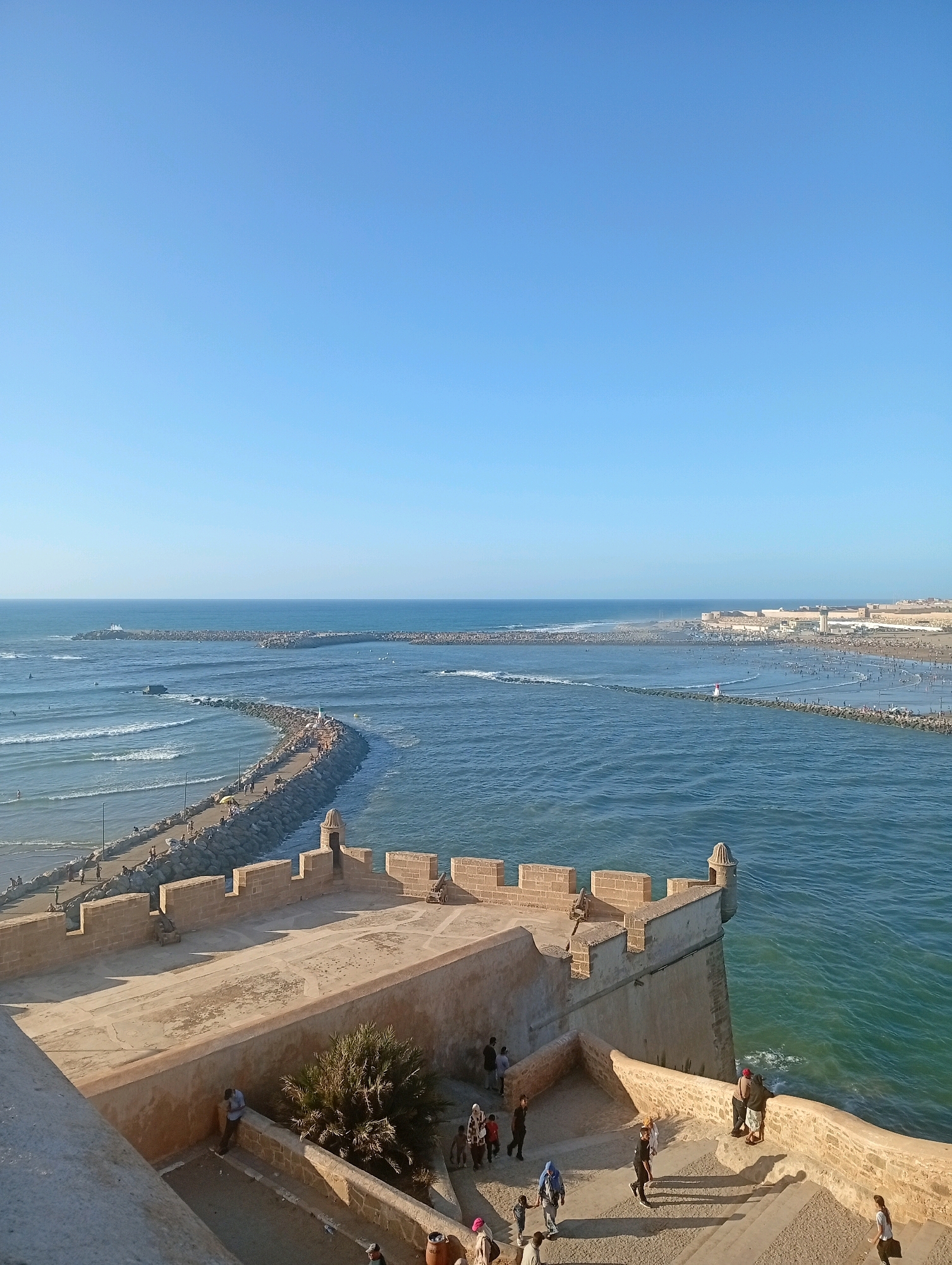 This is an elevated, wide-angle, eye-level shot of the Atlantic Ocean meeting land at the historic Kasbah of the Udayas in Salé, Morocco, during the day. The clear, light blue sky occupies the upper half of the frame. The lower half is dominated by a fortified stone structure, likely a section of the kasbah, with crenellated walls and watchtowers. Several people are visible on the ramparts and walkways of the kasbah, appearing to be tourists or residents. Some are standing, observing the view, while others are walking.
The sea is a vibrant blue-green, with moderate waves breaking and forming whitecaps, particularly near the shore. A long, stone breakwater, also lined with people, curves out into the ocean, creating a protected harbor or channel. Further out, another, larger breakwater extends into the sea, with a town or settlement visible on the coastline in the distance. The beach area adjacent to the breakwaters is crowded with many people enjoying the seaside. The overall scene conveys a sense of a popular tourist destination on a clear, sunny day.