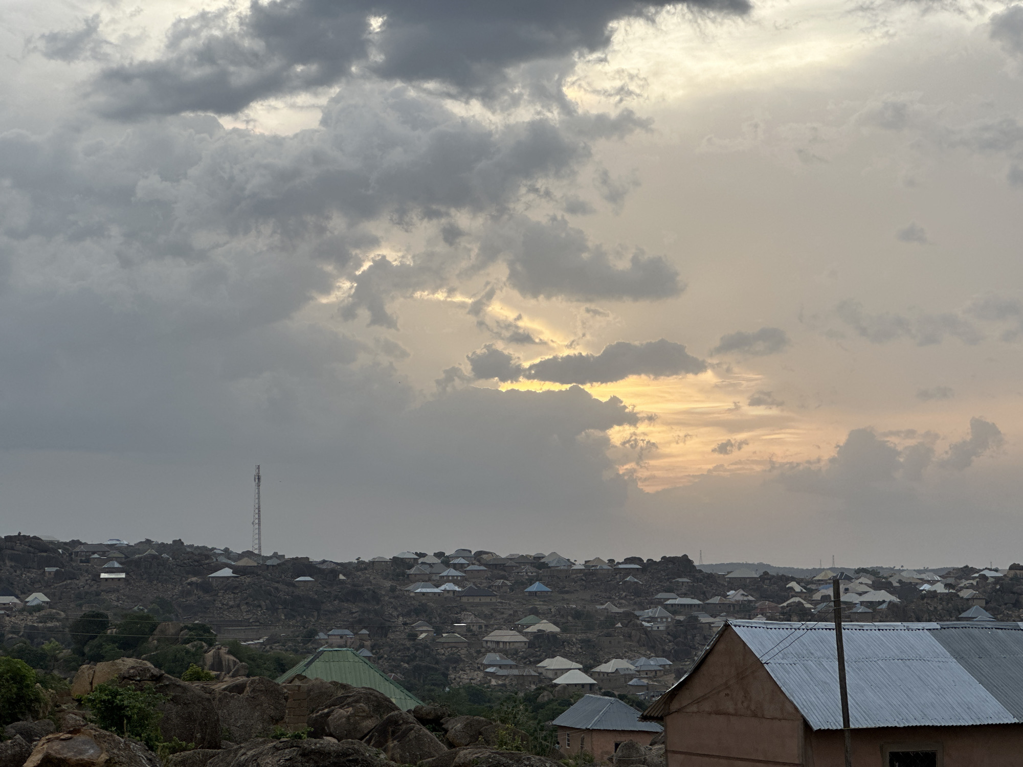 The image depicts a view of the city of Jos, Nigeria, taken during a cloudy sunset. The foreground shows rocky terrain and vegetation. In the middle ground is a densely populated area with houses nestled among the rocks. The roofs of the houses are primarily silver, blue, and green. A tall communication tower stands on a high point in the distance. Above, the sky is filled with dark gray and white clouds, with the setting sun casting a yellow glow through a break in the clouds.