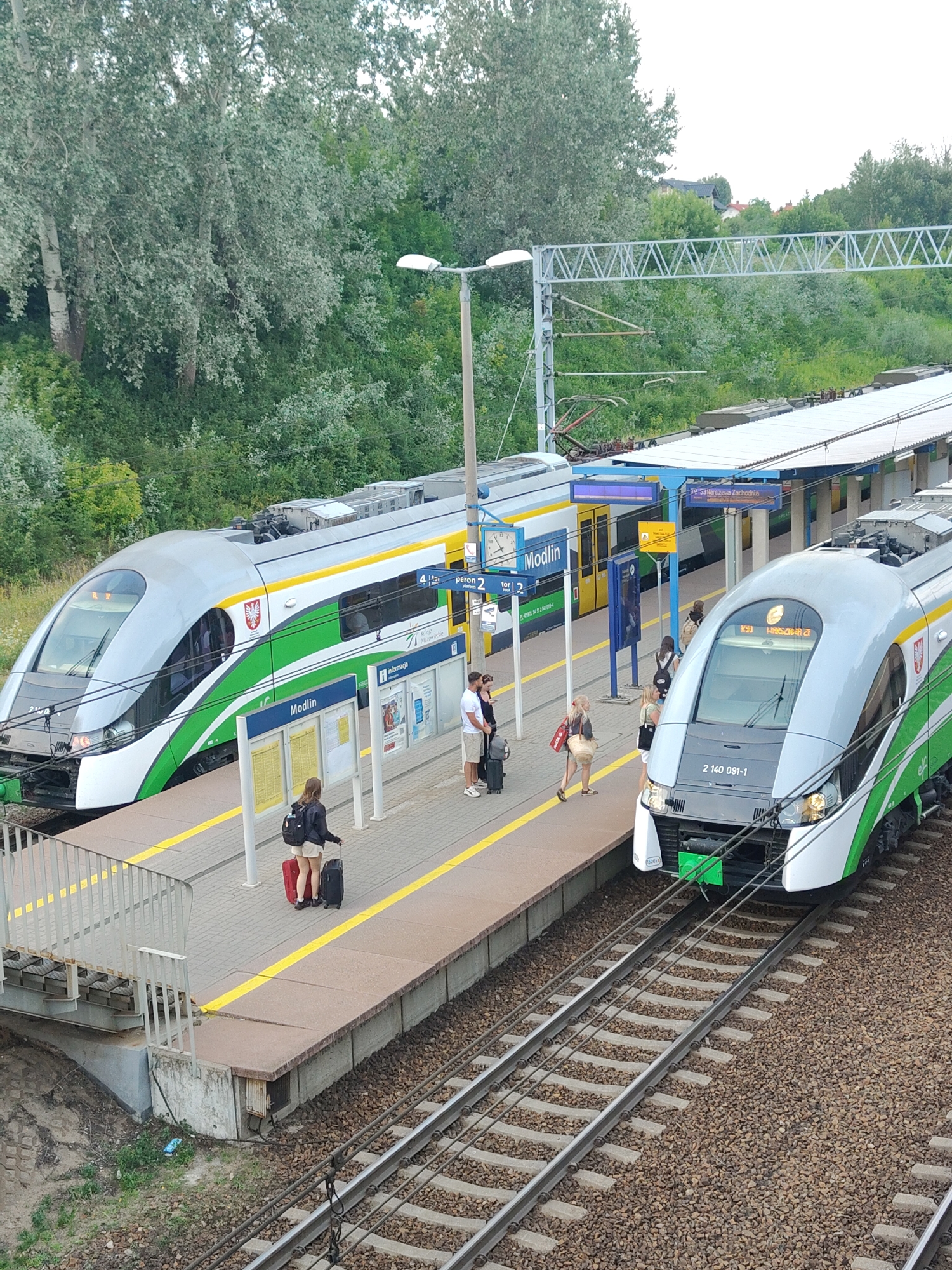 This image captures a train station scene in Nowy Dwór Mazowiecki, Poland. Two modern passenger trains, one silver with green and white accents and the other predominantly silver with green stripes, are visible on adjacent tracks. The train on the left has the word "Modlin" displayed prominently, indicating its destination or route. Several people are on the platform, some with luggage, suggesting they are either waiting to board or have just disembarked.
The platform itself is paved and marked with yellow safety lines. Information boards are placed along the platform, providing schedules and details about train services. One board clearly shows "Modlin" and another refers to "peron 2 tor 2" (platform 2 track 2). Further signage visible includes "Rinformaça" and "19:33 Warszawa Zachodnia", indicating a train departing for Warsaw West at 19:33.
The background features lush green trees and vegetation, hinting at a somewhat rural or suburban setting. Overhangs from a station roof provide shelter. The lighting suggests it's late afternoon or early evening, with diffused natural light. The overall atmosphere appears calm and functional, typical of a train station environment. The trains are sleek and modern, equipped with overhead power lines. The text "2 140 091-1" is visible on the front of the train on the right.