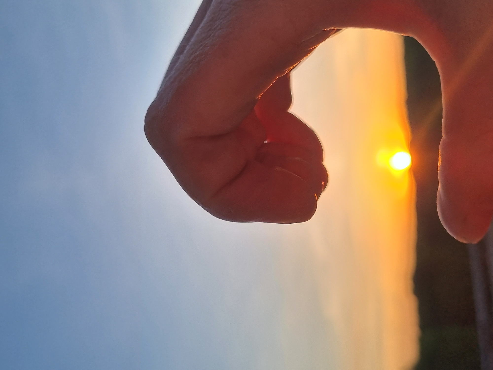 A human hand is positioned against a sky transitioning from blue to orange, with the setting sun partially obscured by the hand. The background suggests a landscape with possible trees or vegetation on the horizon. The photograph does not provide sufficient information to ascertain the location in Kemul, Russia.