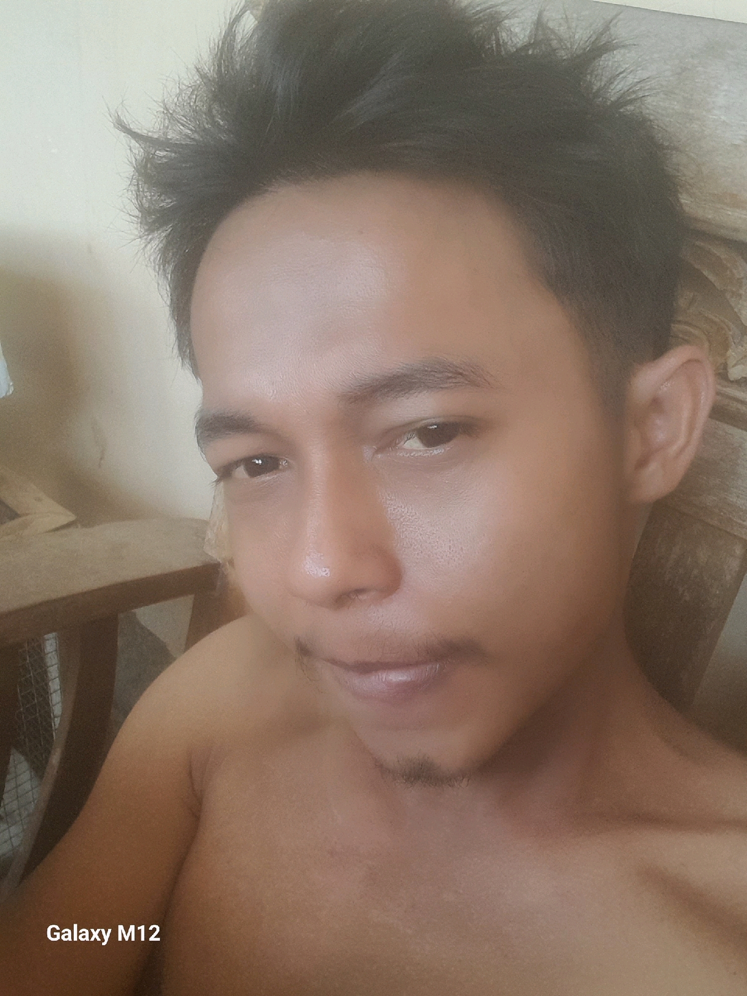 The image is a selfie taken by a young man in Karawang, Indonesia. The man has dark, spiky hair and is shirtless. He is looking directly at the camera with a neutral expression. The background appears to be an indoor setting with a light-colored wall and a wooden chair visible. The lower left corner of the image contains the text "Galaxy M12," indicating the device used to take the photo.